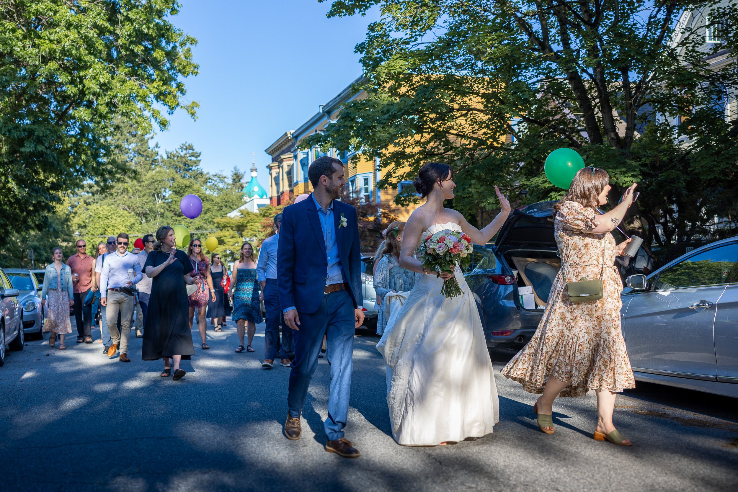 A bride in a white wedding dress holding a bouquet, walking alongside a groom in a blue suit, as part of a wedding procession through a street lined with parked cars and guests, with balloons and trees in the background.