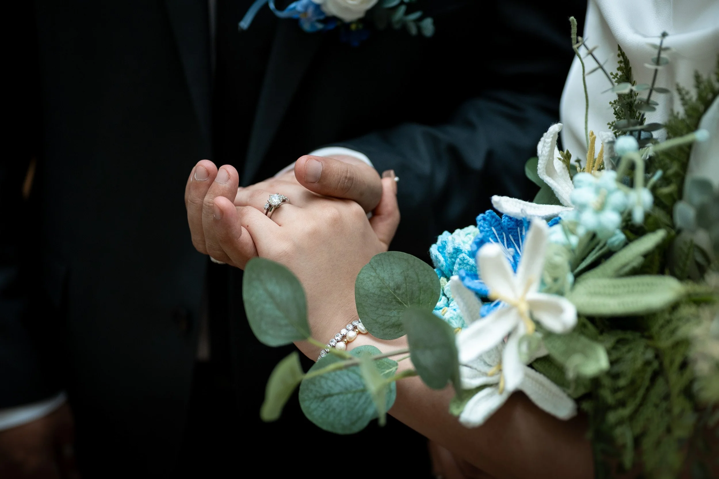 Close-up of a couple holding hands, with a focus on an engagement ring on the woman's finger, surrounded by a bouquet of flowers.