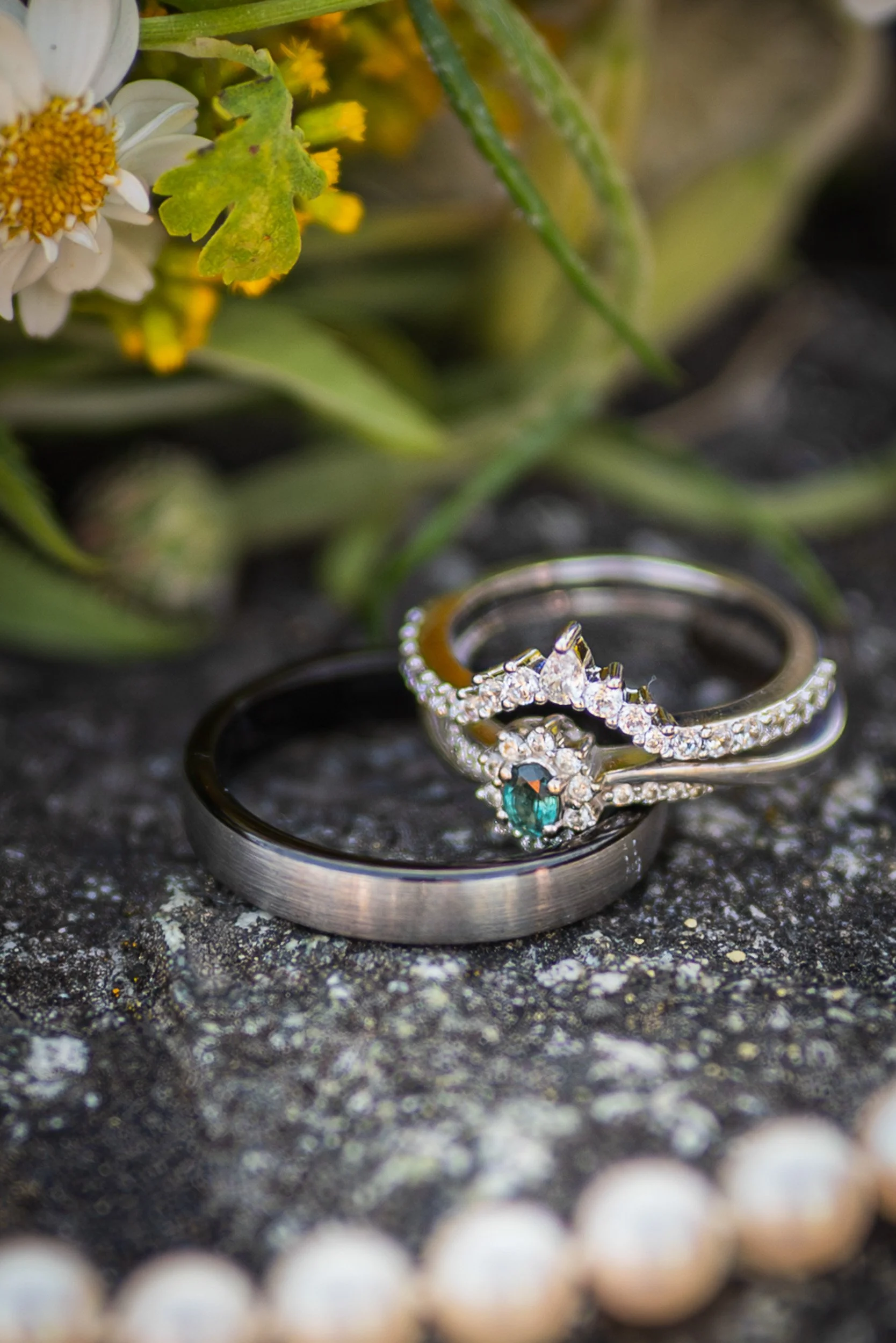 Close-up of a diamond engagement ring and wedding band on a textured surface, with blurred white beads in the foreground and some yellow flowers and green leaves in the background.