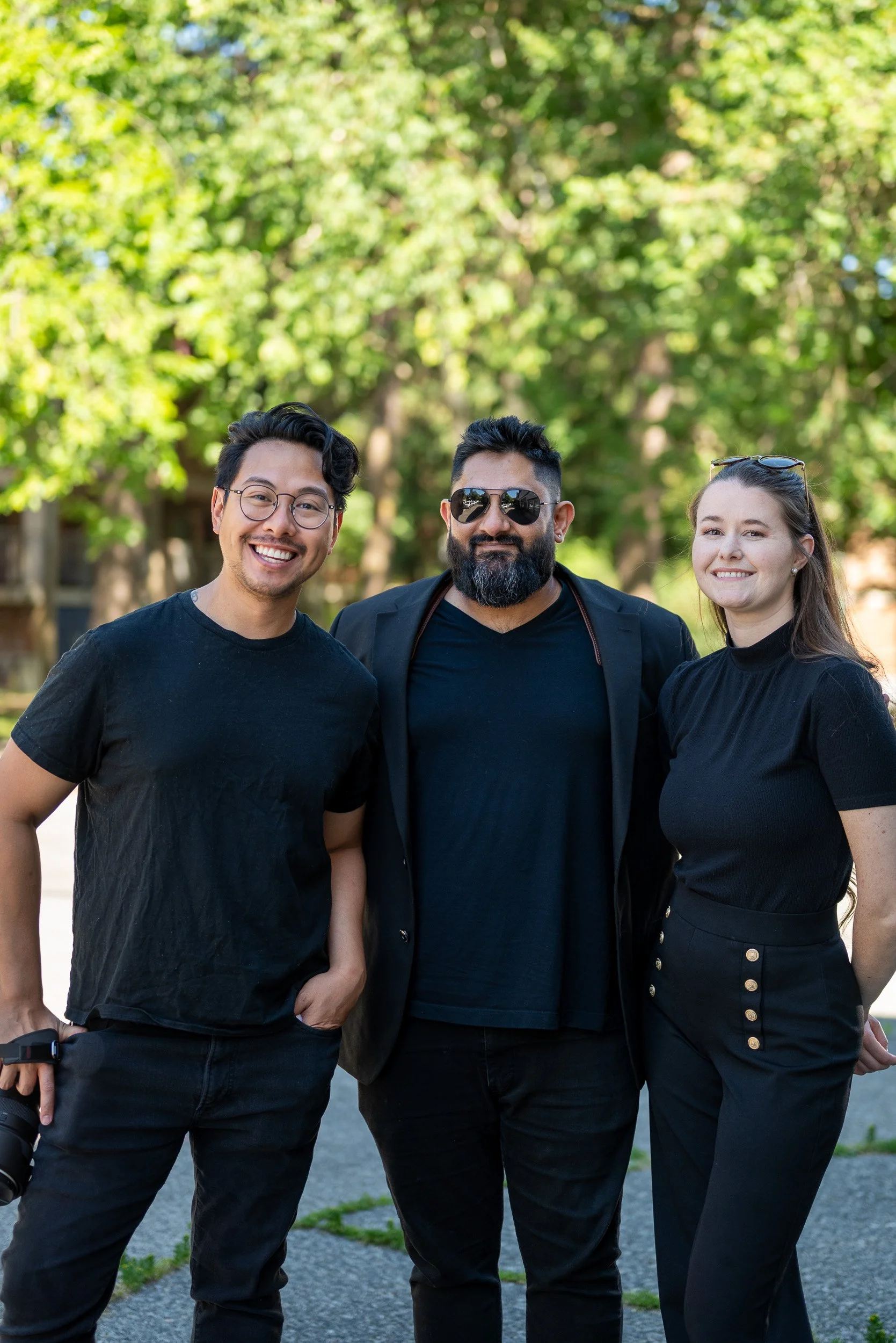 Three people standing outdoors in front of green trees, smiling at the camera. The person on the left has glasses and curly hair, the middle person has sunglasses and a beard, and the right person has glasses on her head and long hair.