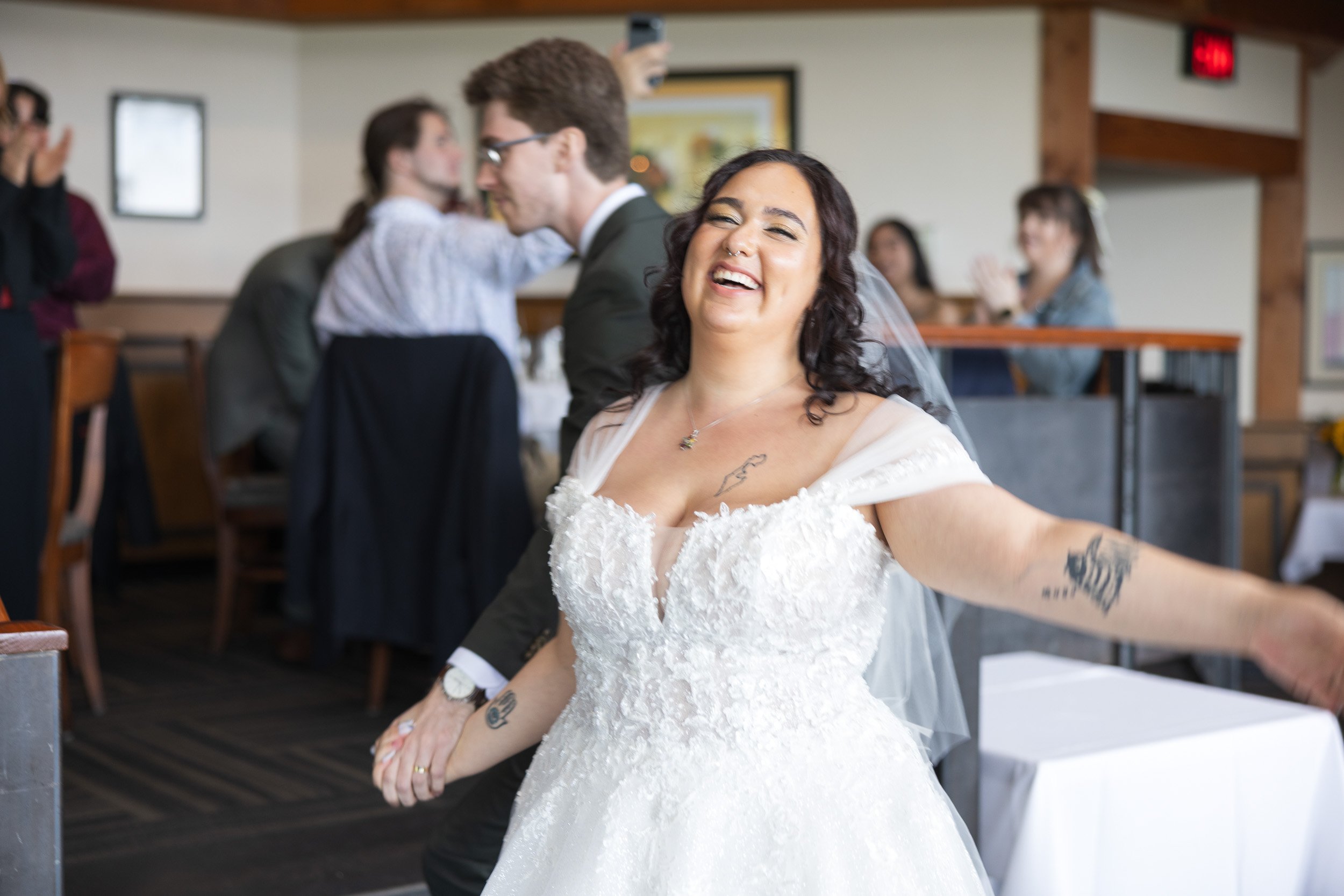 A joyful bride with dark curly hair, tattoos, and a nose ring, wearing a white wedding dress, holding hands with her groom, in a reception hall with seated guests clapping and watching in the background.