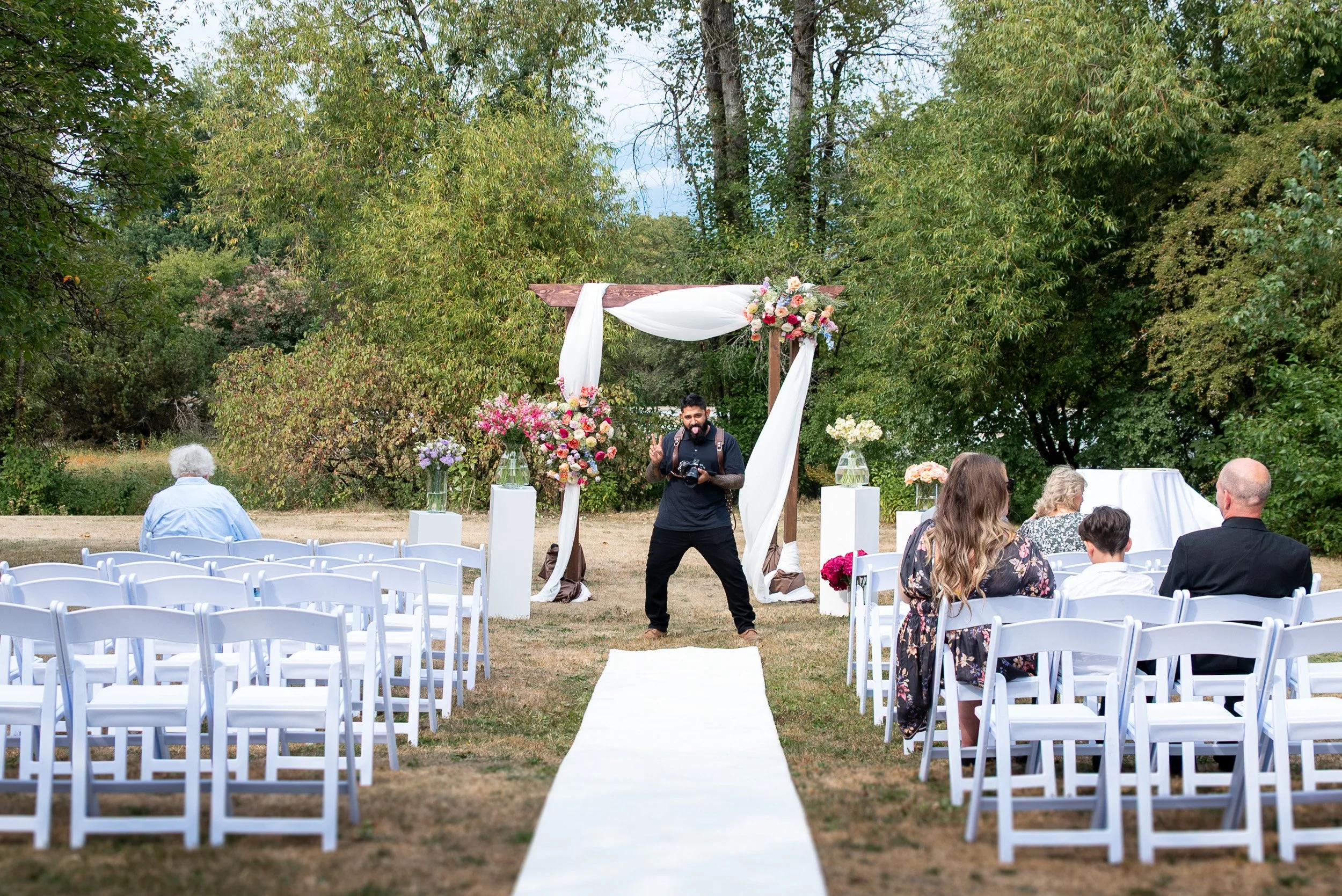 Outdoor wedding ceremony setup with white chairs arranged in rows, a white aisle runner, a floral arch with pink and white flowers, and guests seated under green trees.