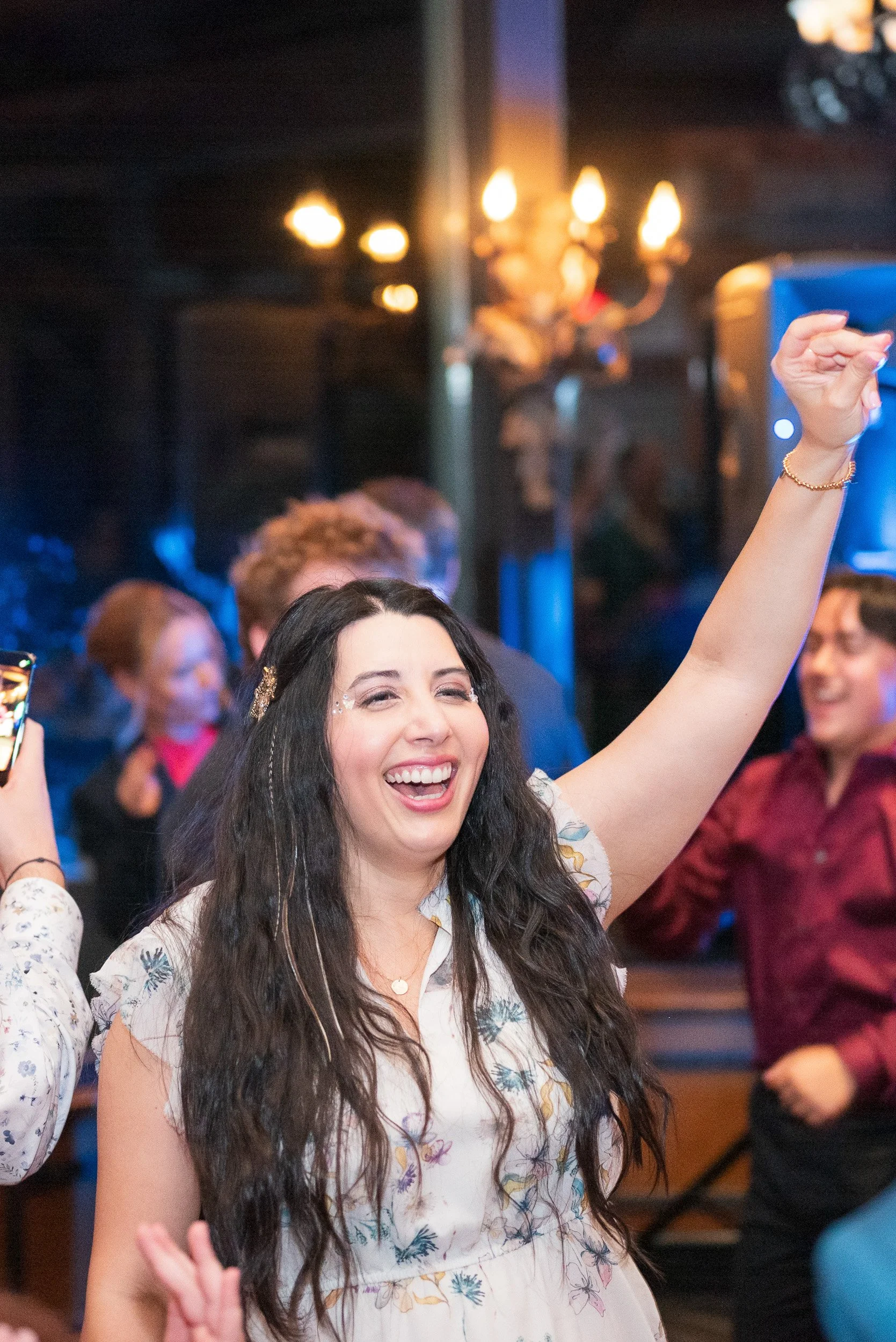 A woman with long dark hair and an expressive face, smiling and raising her arm in a lively indoor setting with warm lighting and other people in the background.