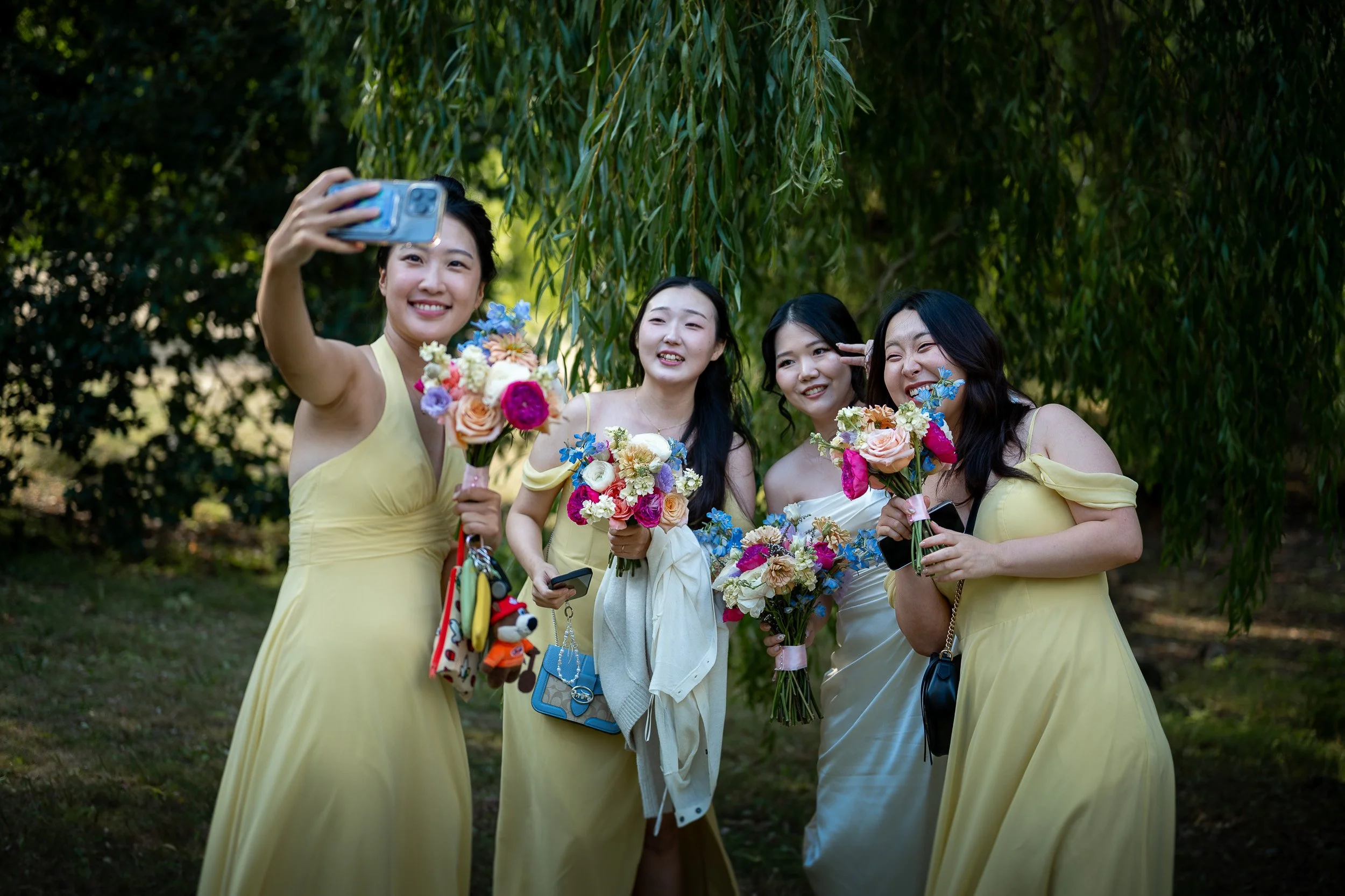 Four women in yellow and white dresses hold bouquets of flowers and take a selfie outdoors with a woman in white in the center. They are smiling and standing under a tree with green leaves.
