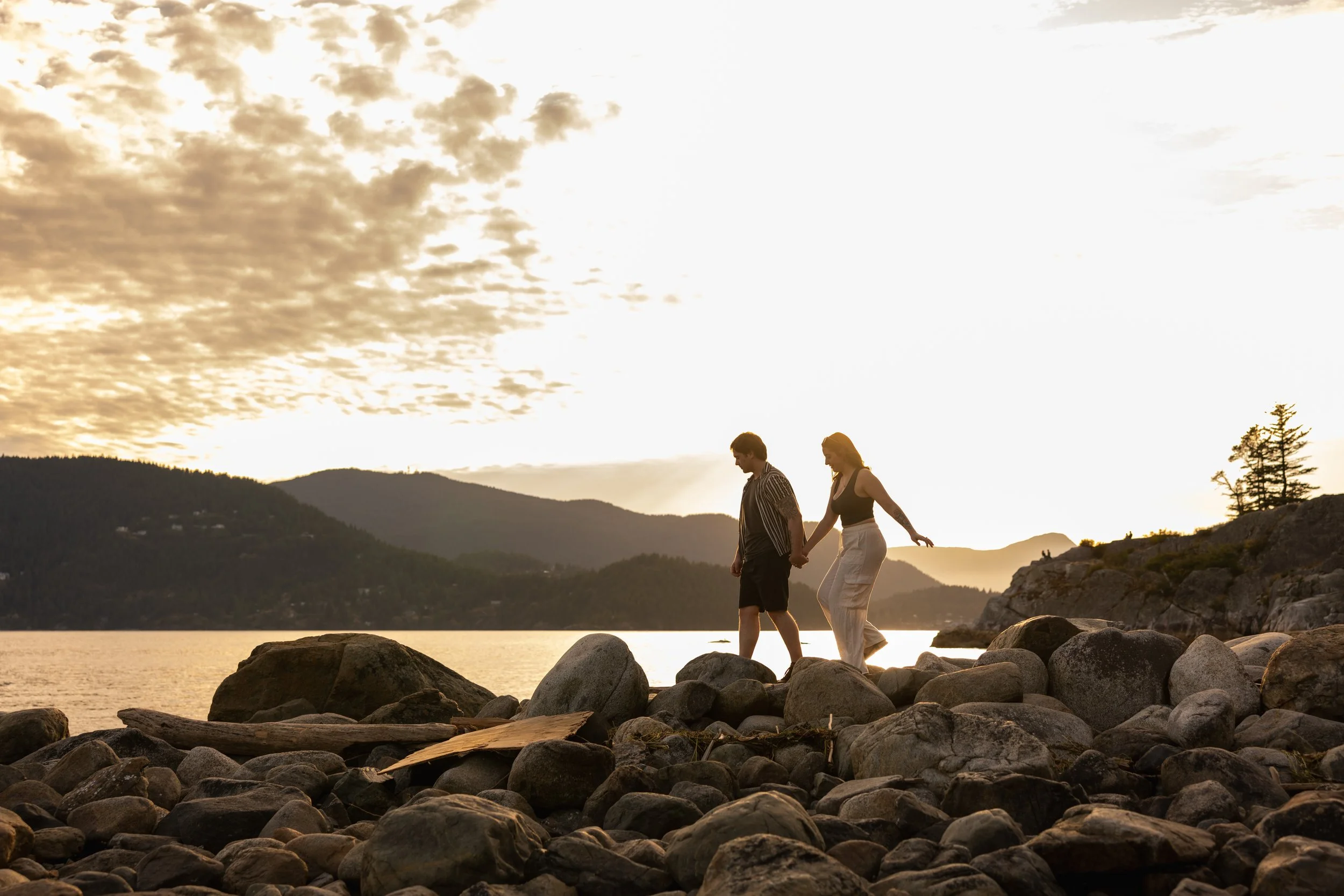 A couple walking hand-in-hand along a rocky shoreline during sunset, with mountains and a partly cloudy sky in the background at Whytecliff Park in West Vancouver.
