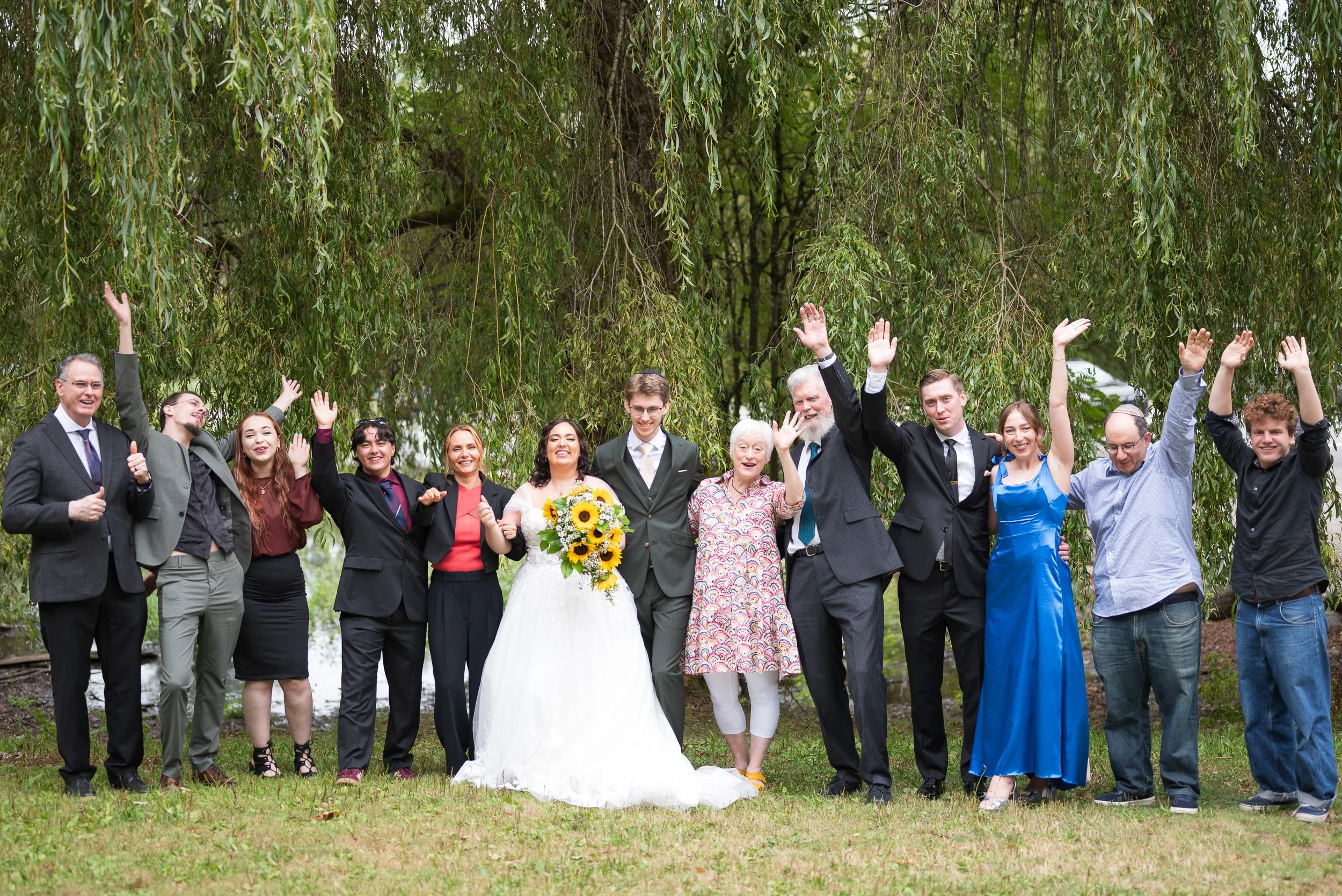 Group of people celebrating a wedding outdoors under a large tree, smiling, waving, with the bride holding a bouquet of sunflowers.