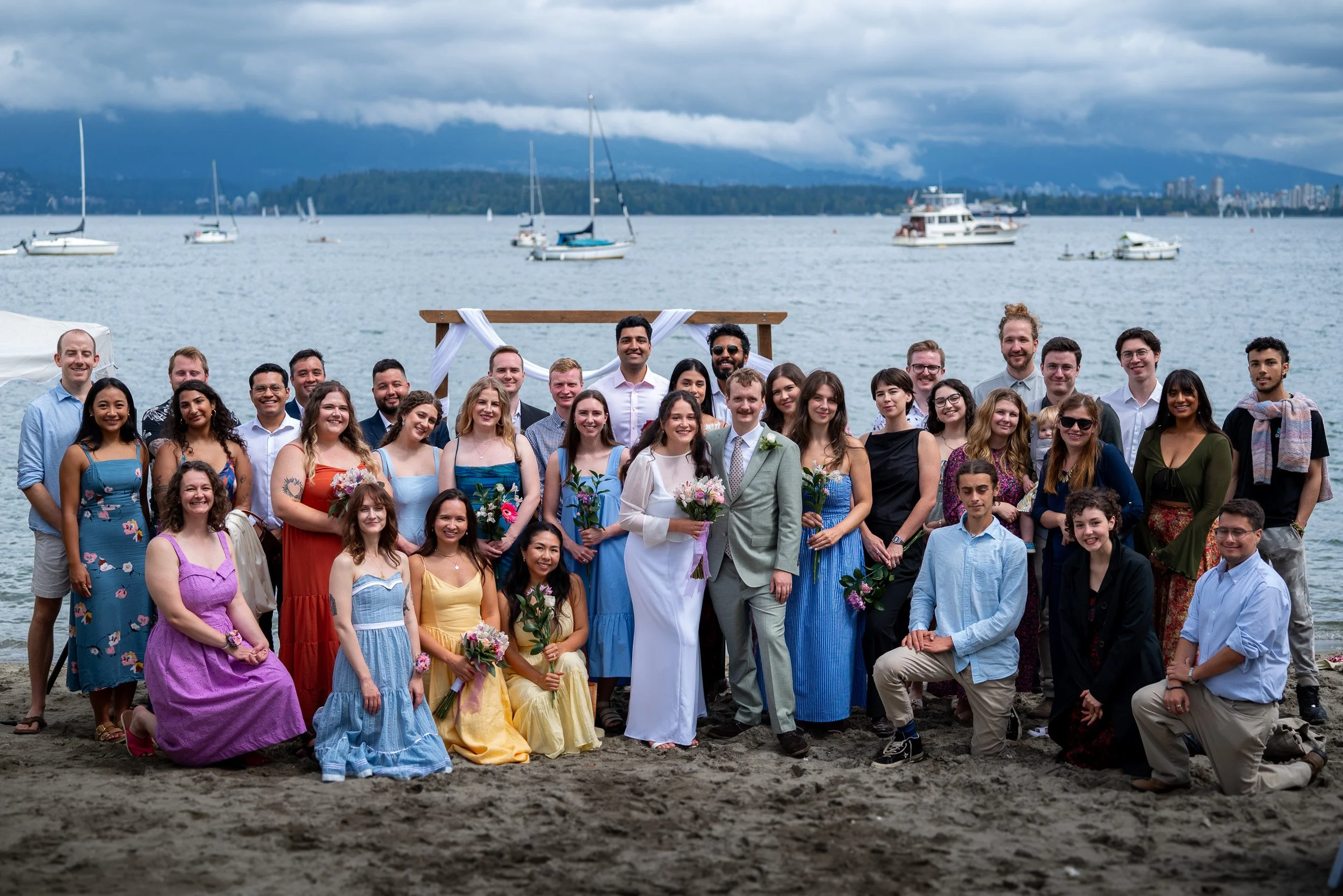 A large group of people gathered on a beach for a wedding celebration at Jericho Beach, with boats and a body of water in the background, under cloudy skies.