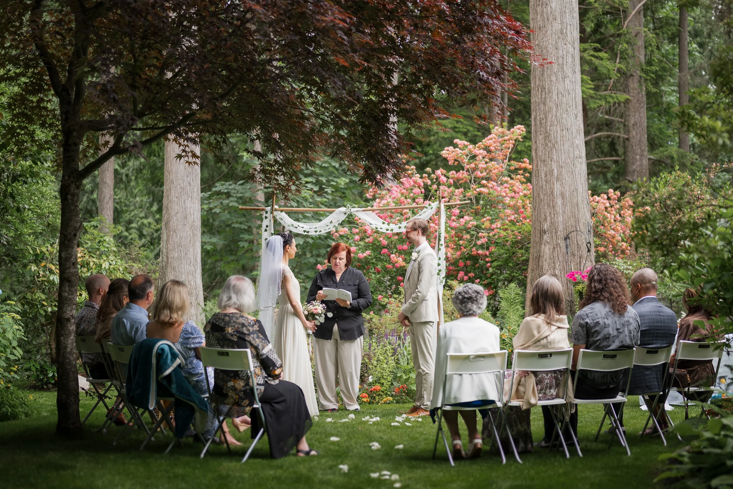 An outdoor wedding ceremony in a lush green garden with tall trees and pink flowers. A bride and groom are standing before an officiant, holding hands, at their micro-wedding in Vancouver
