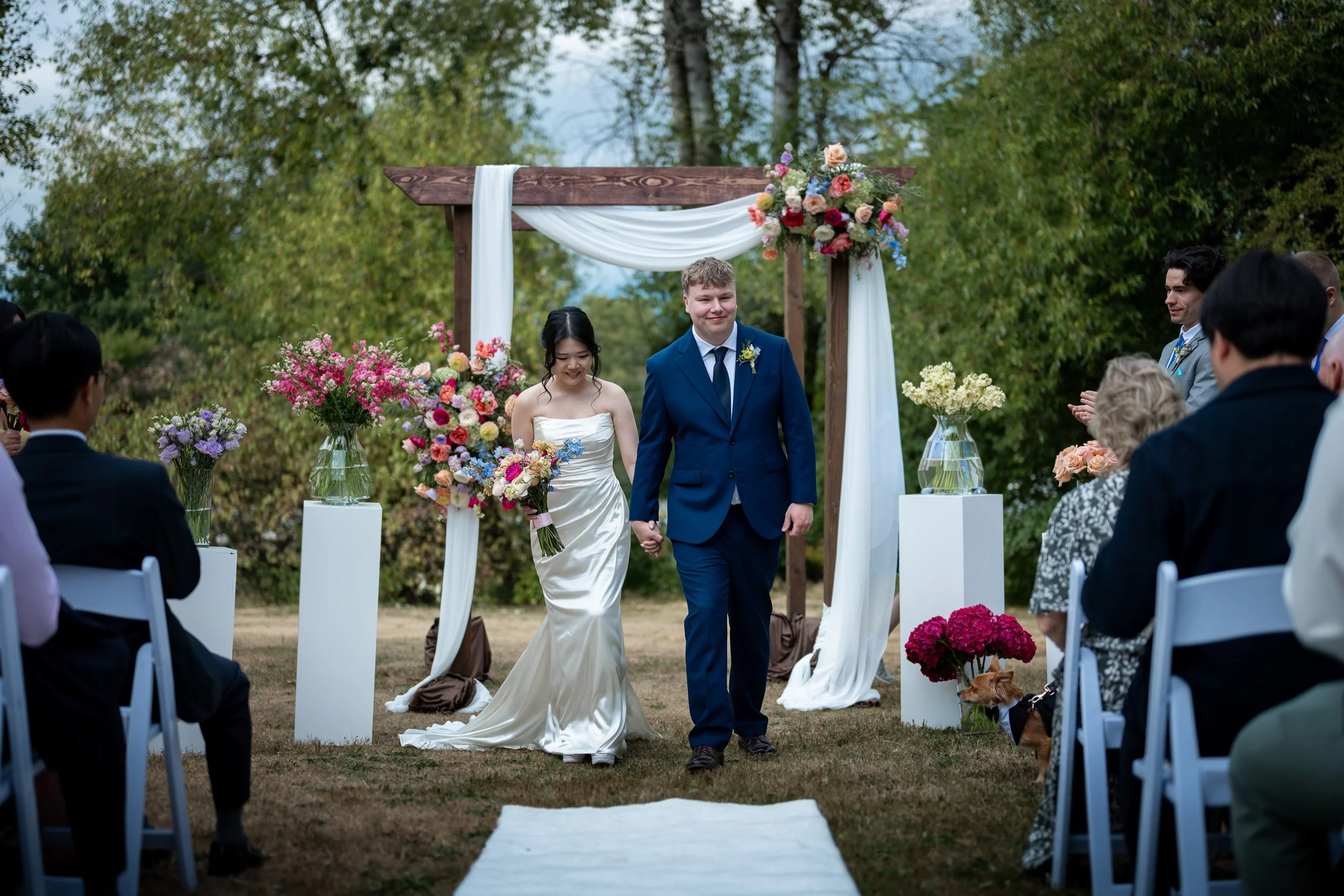A bride and groom holding hands walking down the aisle during an outdoor wedding ceremony surrounded by guests and decorated with flowers and drapery under a wooden arch at Queen Elizabeth Park.