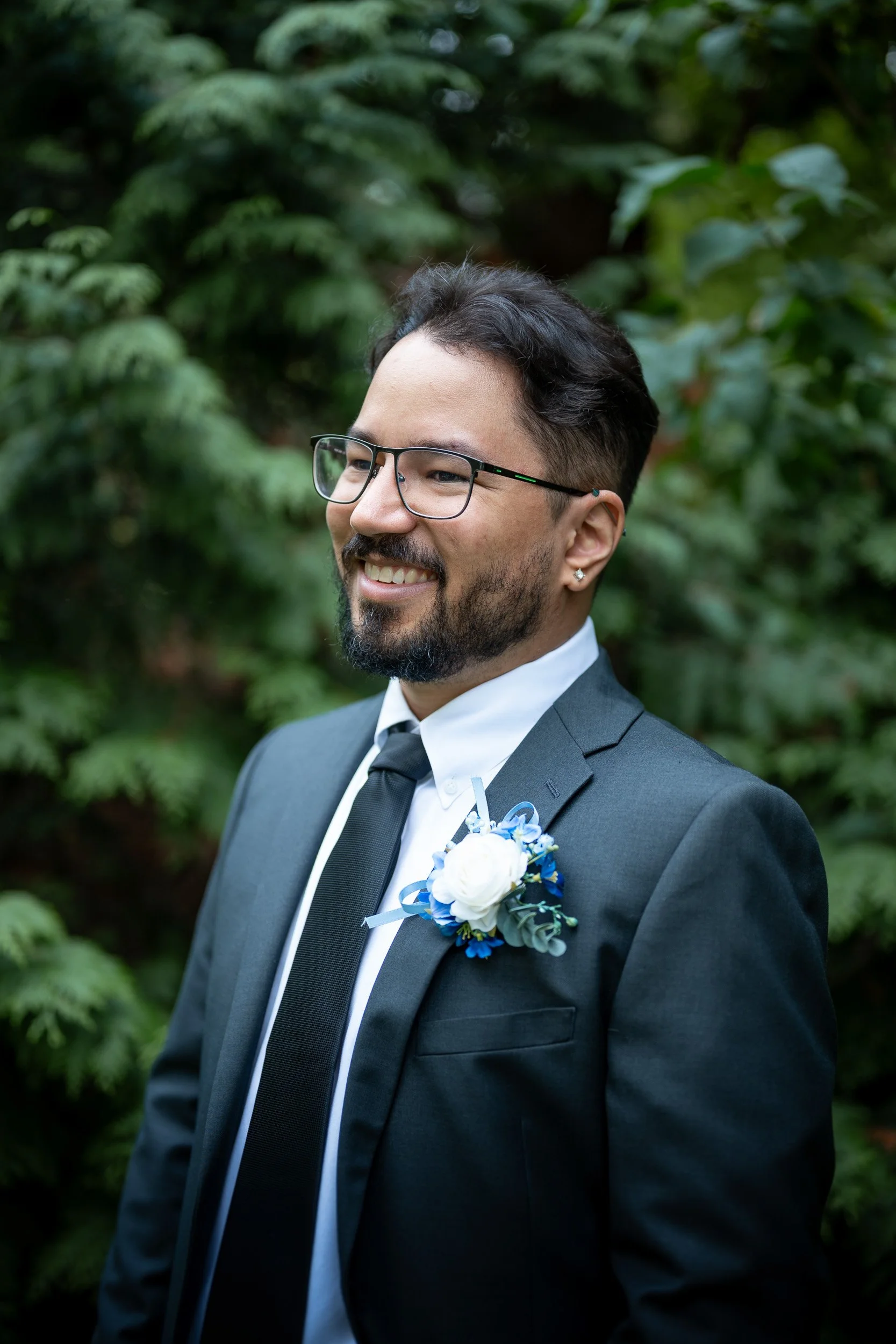Close-up of a smiling man with glasses and a beard in a suit with a boutonniere, standing outdoors with green foliage in the background.