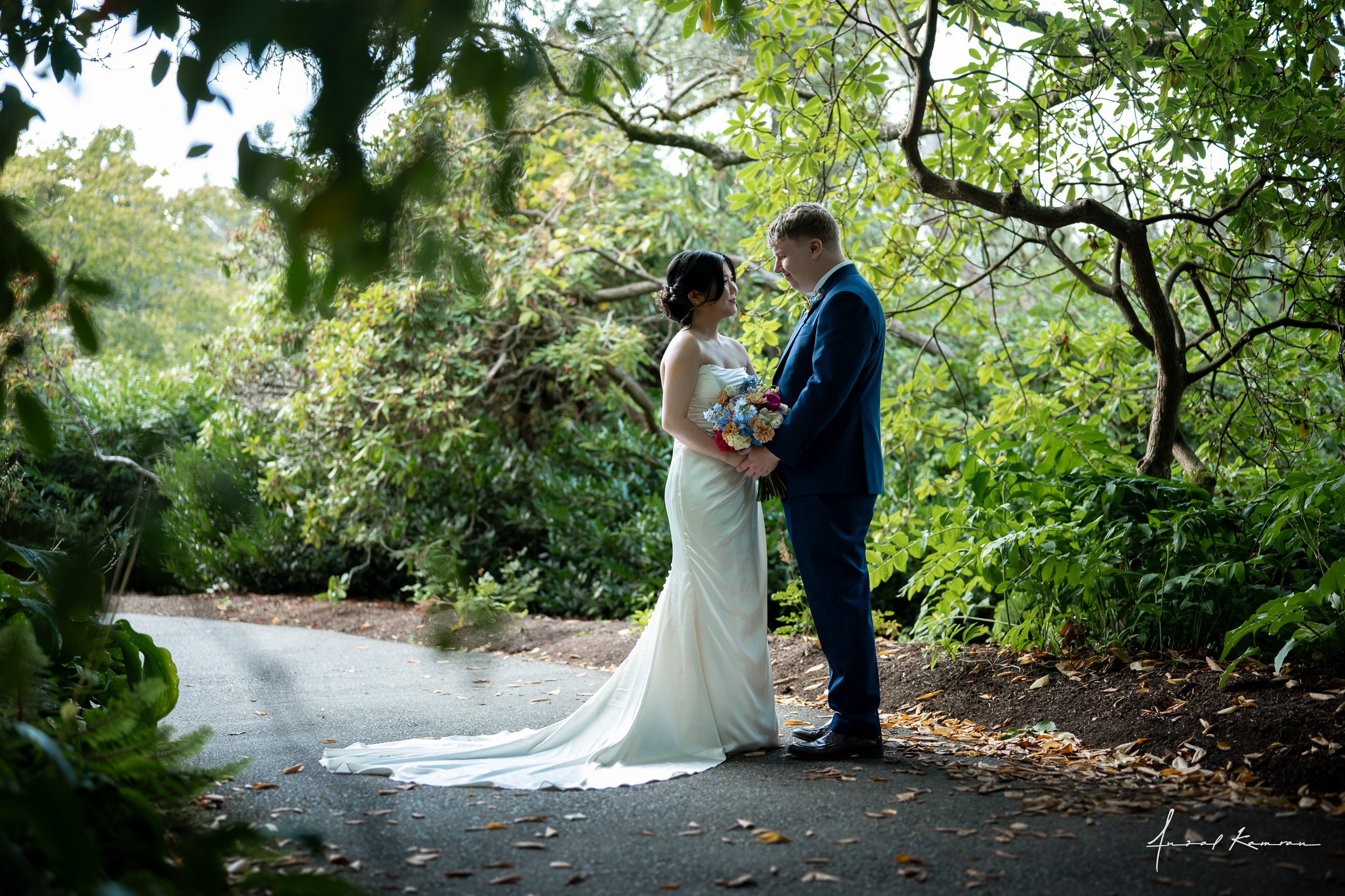 A bride and groom stand facing each other on a paved path surrounded by lush green trees, with the bride holding a bouquet of flowers and wearing a strapless wedding gown, while the groom is dressed in a blue suit at Queen Elizabeth Park.