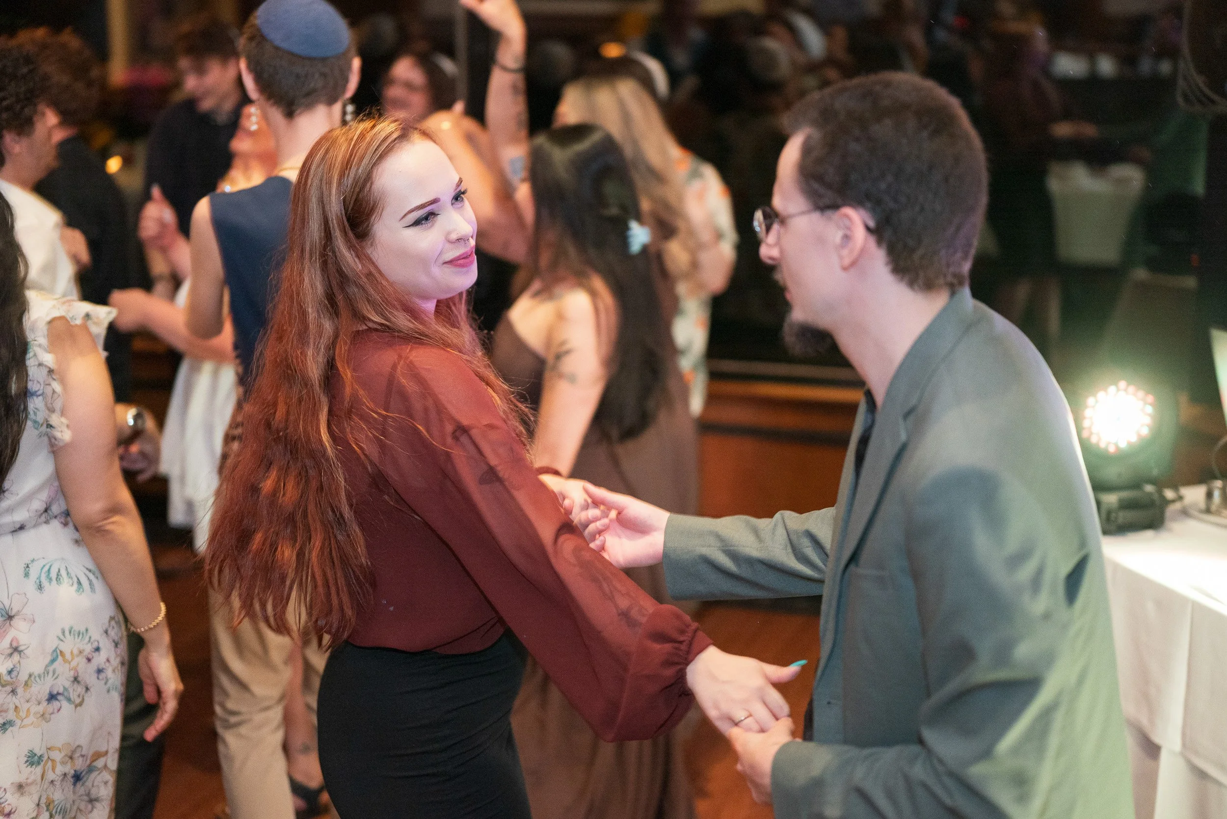 A woman with red hair and a septum piercing shaking hands with a man wearing glasses and a gray suit at a social event. Several people are in the background, some in conversation, in a dimly lit indoor setting.