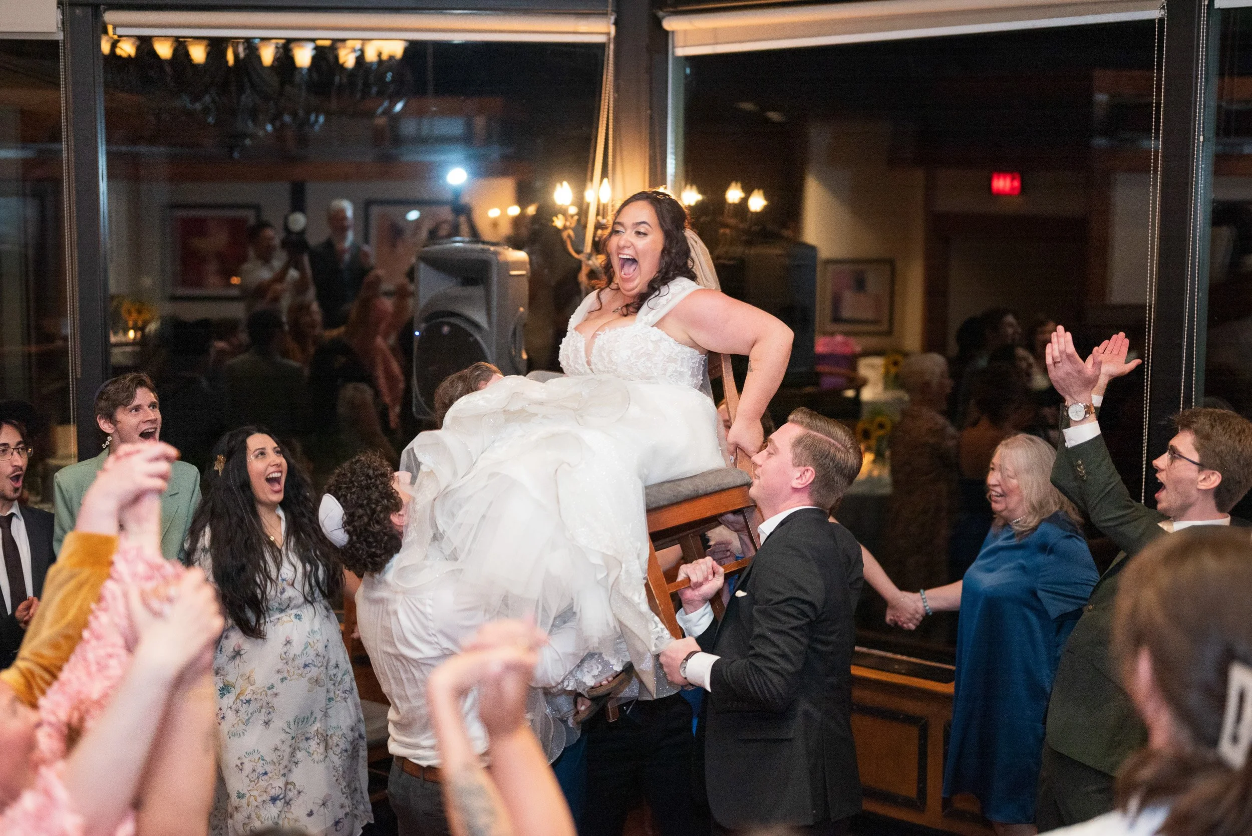 A bride in a wedding dress is being lifted by multiple people during a wedding celebration, surrounded by joyful guests clapping and cheering.