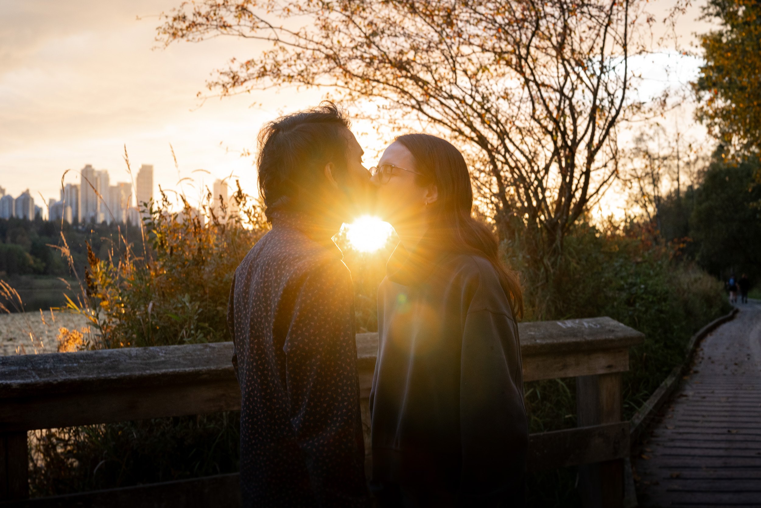 A couple is kissing against the sunset in a park with autumn trees and city skyline in the background.