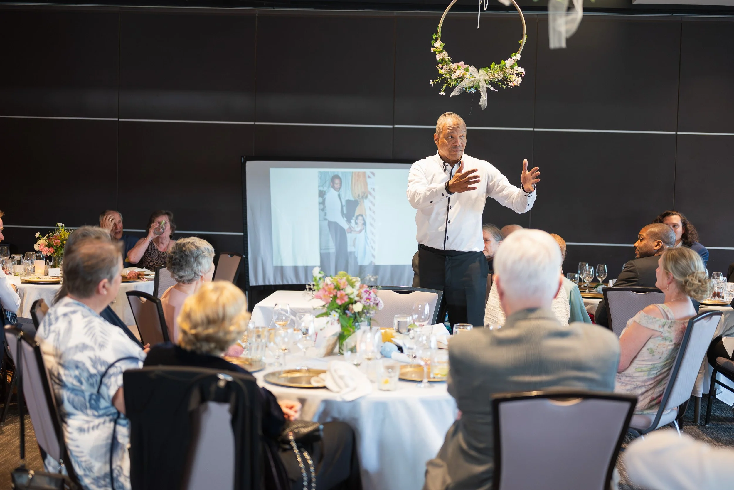 A man in a white shirt giving a speech at a wedding at Seymour Golf and Country Club. The audience is seated at round tables decorated with floral centerpieces.
