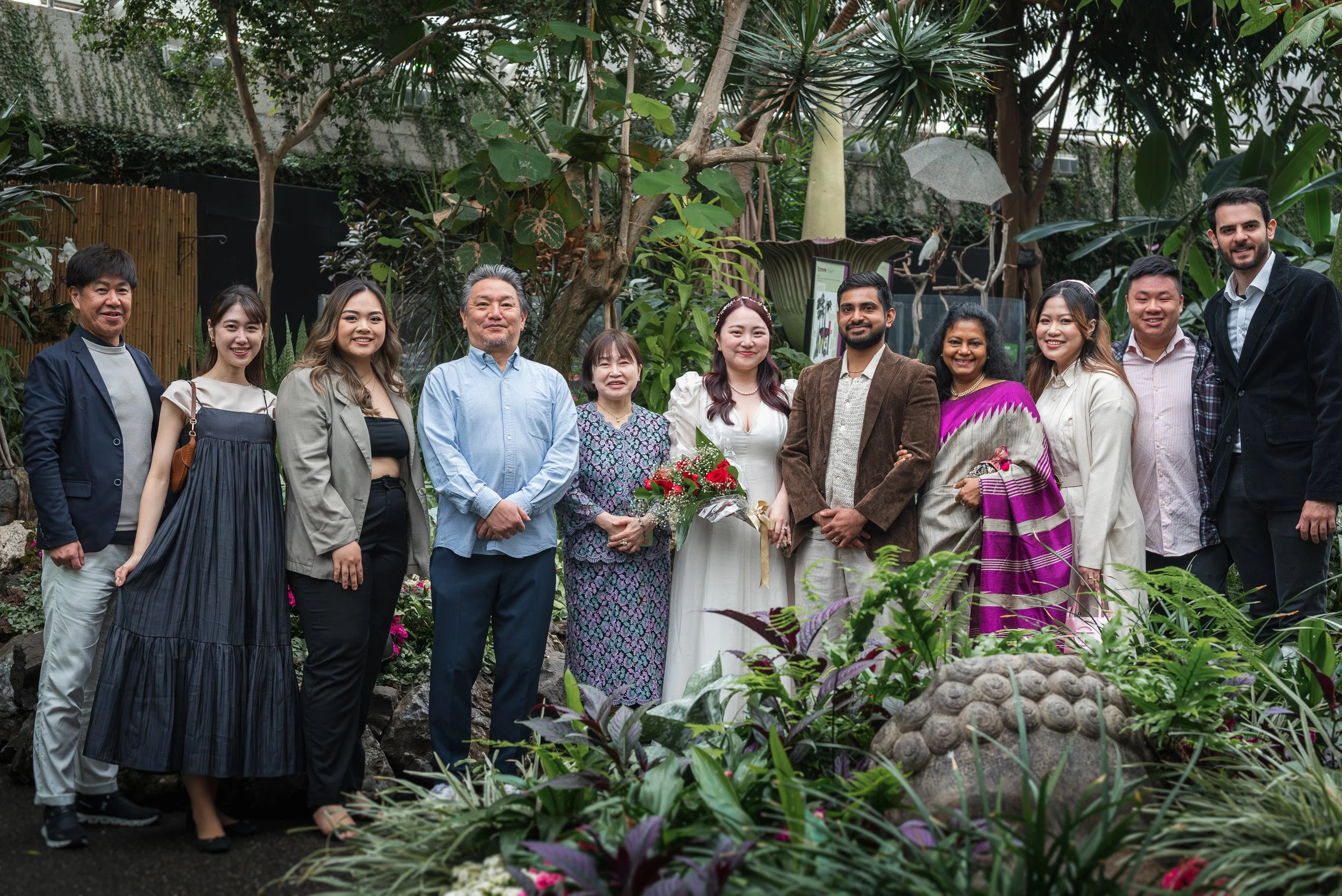 A diverse group of eleven people standing together in a lush indoor garden, smiling for a photo, with tropical plants and trees in the background.