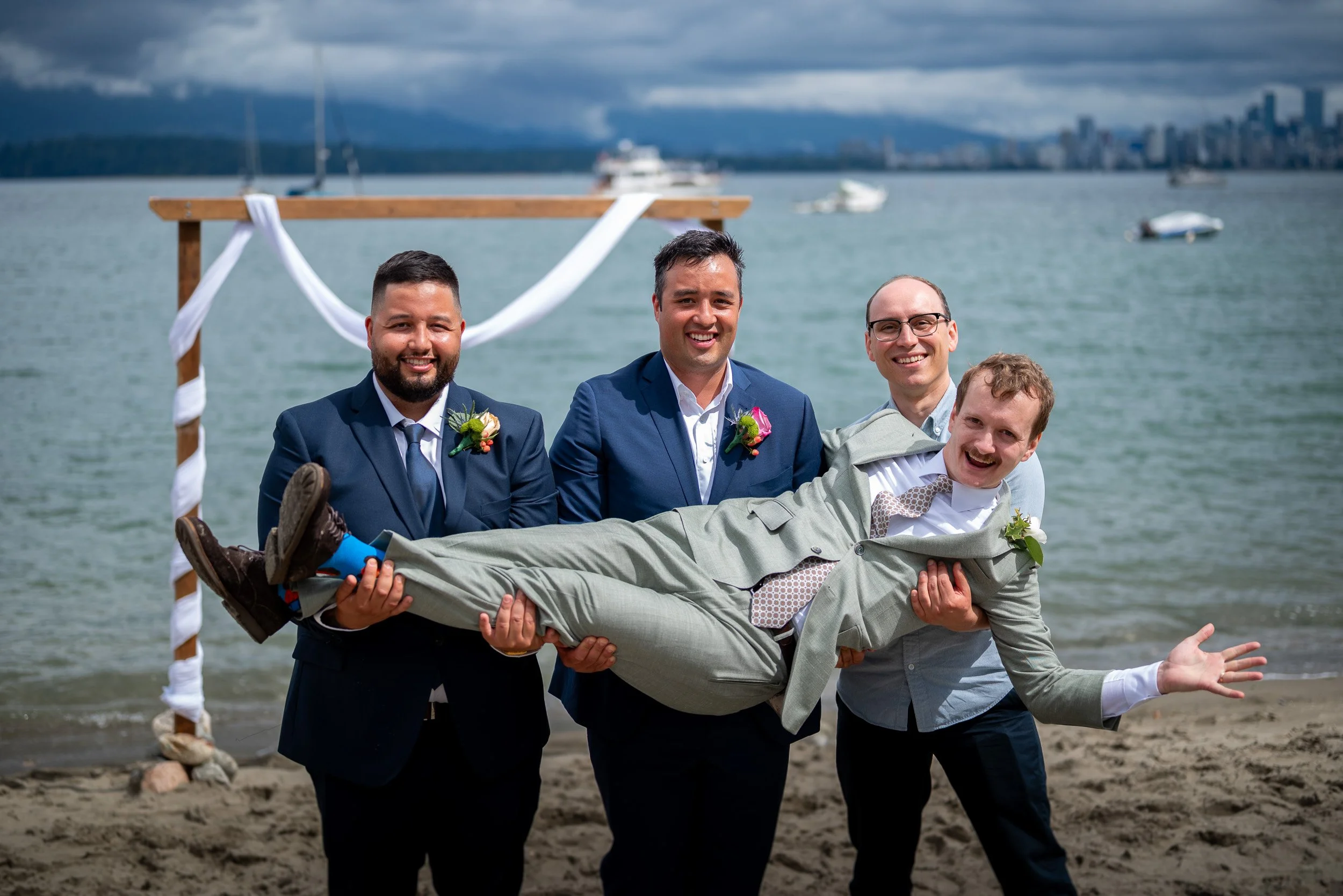 Four men in suits and dress shirts, smiling, holding a fifth man horizontally in front of them at a beach with boats and city skyline in the background.
