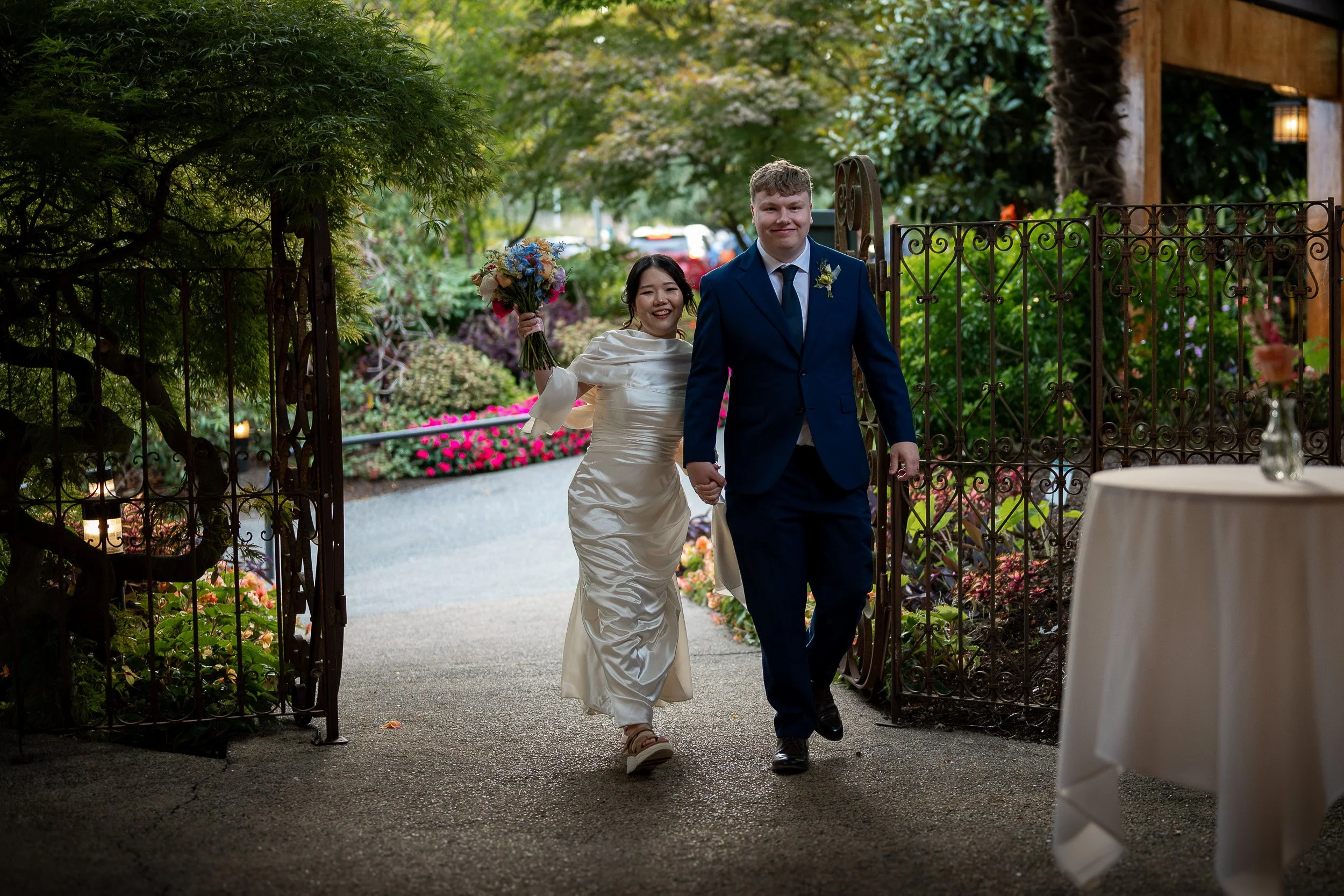 A bride and groom walking hand in hand through the entrance of Season in the Park Restaurant, smiling. The bride is holding a bouquet and wearing a white satin dress. The groom is dressed in a blue suit with a white shirt and dark tie.