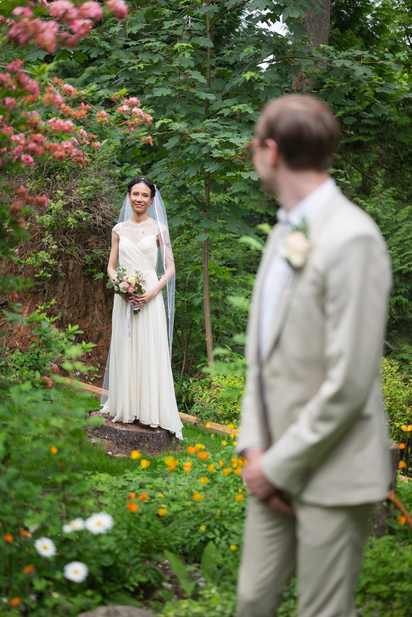A bride in a white wedding dress and veil standing in a lush green garden, holding a bouquet of flowers, looking at a groom in a light-colored suit with glasses, who is out of focus in the foreground.