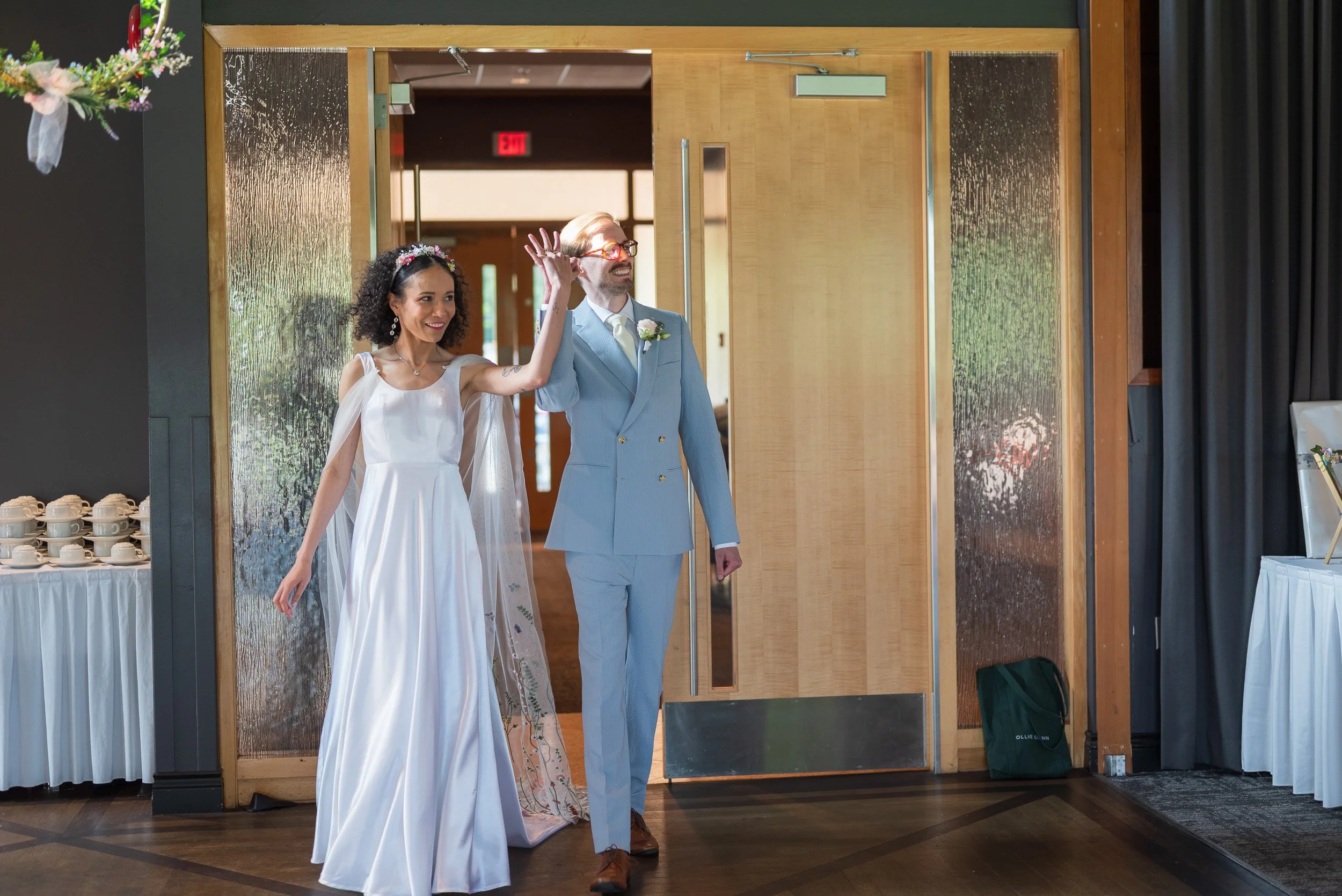A bride and groom entering the Seymour Golf and Country Club hand in hand. The bride has curly hair and a floral headband, wearing a white wedding dress. The groom has blond hair, glasses, and a light blue suit. They are smiling as they walk inside.