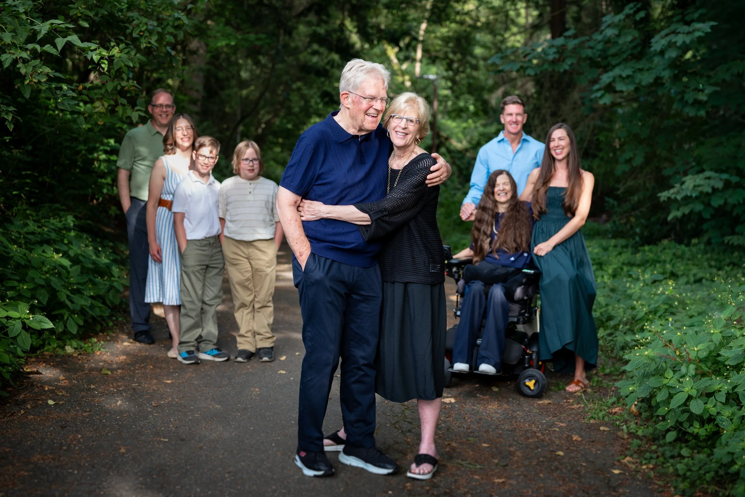 A group of family members smiling and hugging in a forested park, with a senior couple in the foreground and children and young adults in the background.