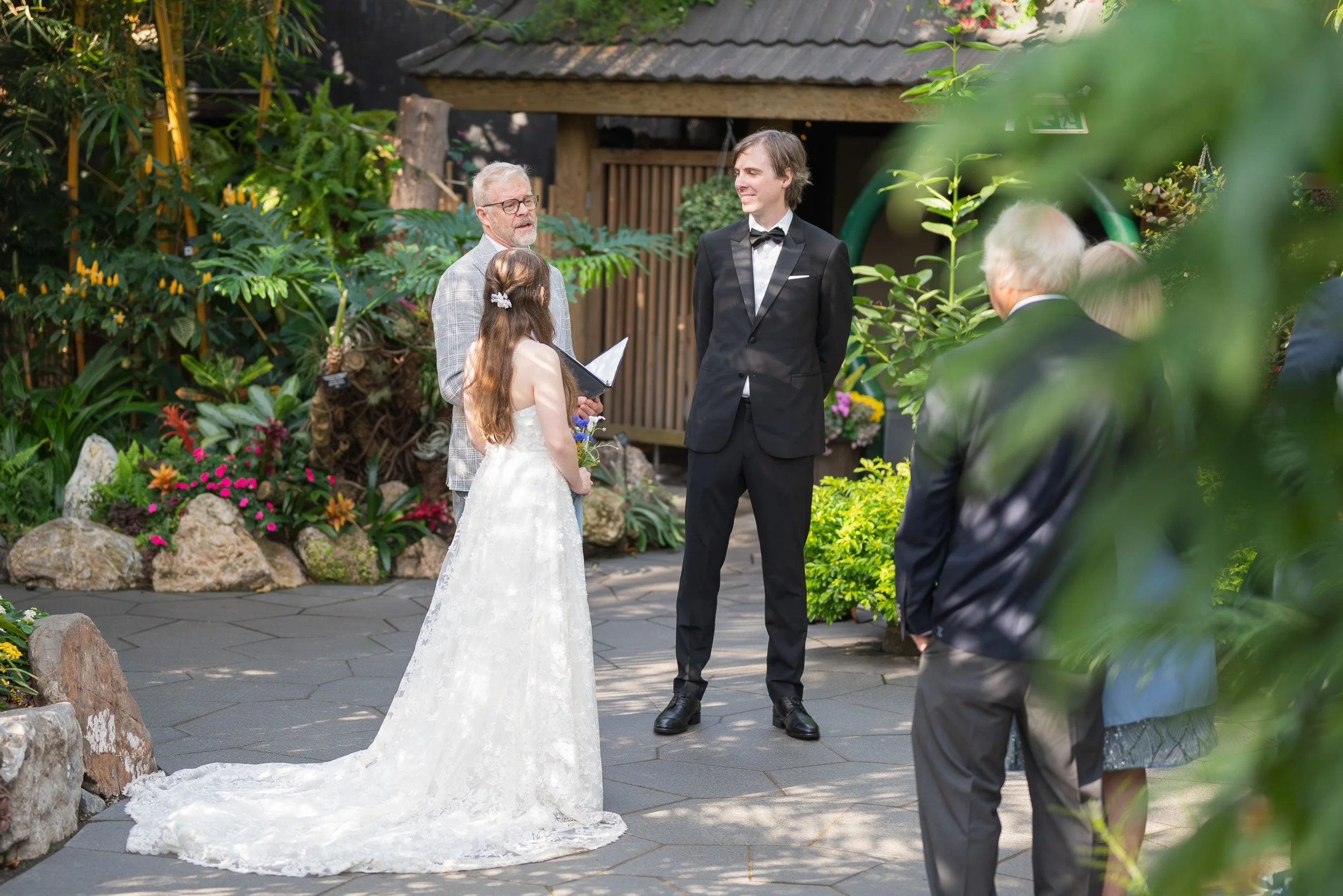 A wedding ceremony taking place at Bloedel Conservatory with a bride in a white dress, a groom in a black tuxedo, and officiants and guests surrounding them amid green foliage and flowers.