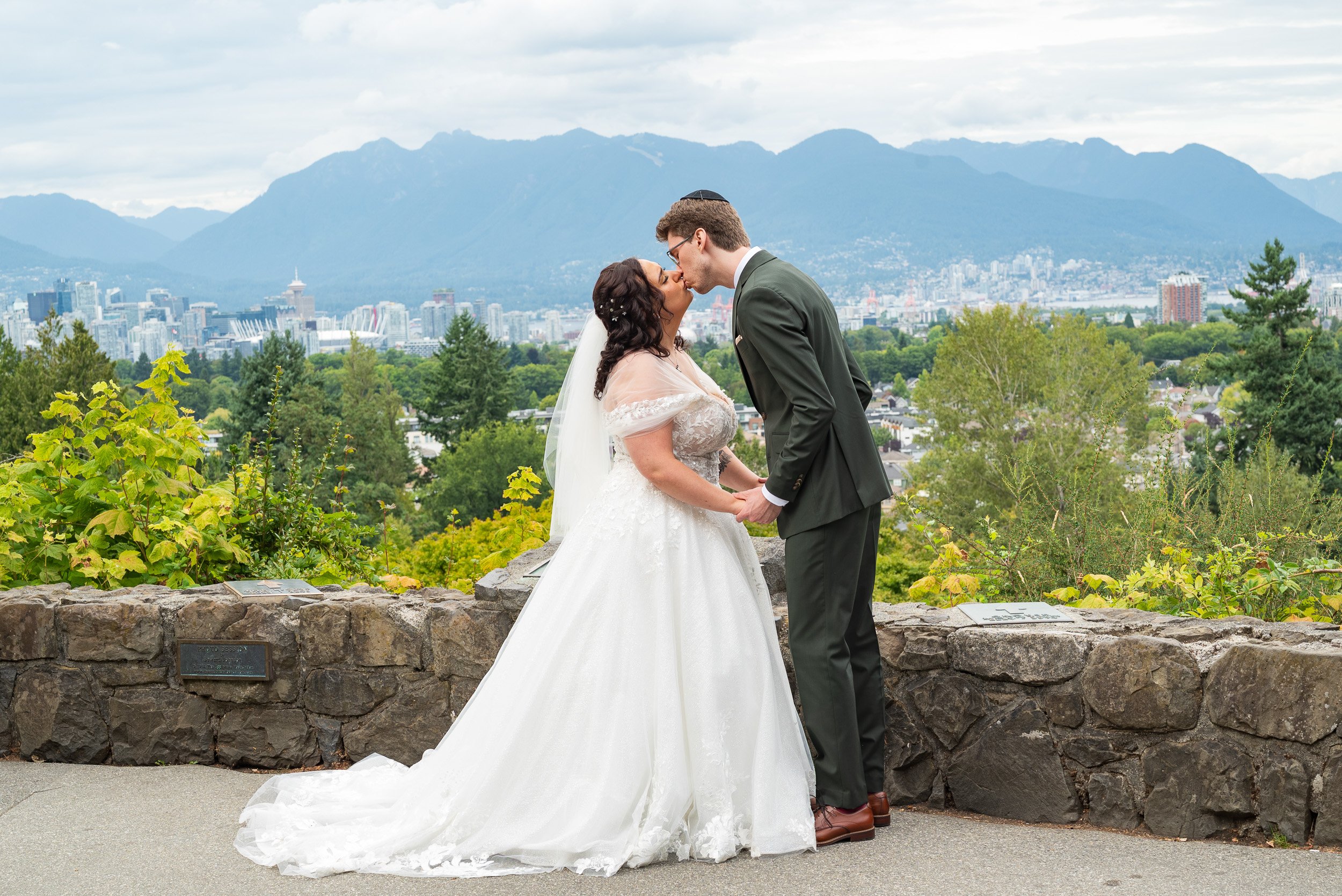 A bride and groom share a kiss during their wedding ceremony outdoors with a Vancouver city skyline and mountains in the background. This is the view from Queen Elizabeth Park.