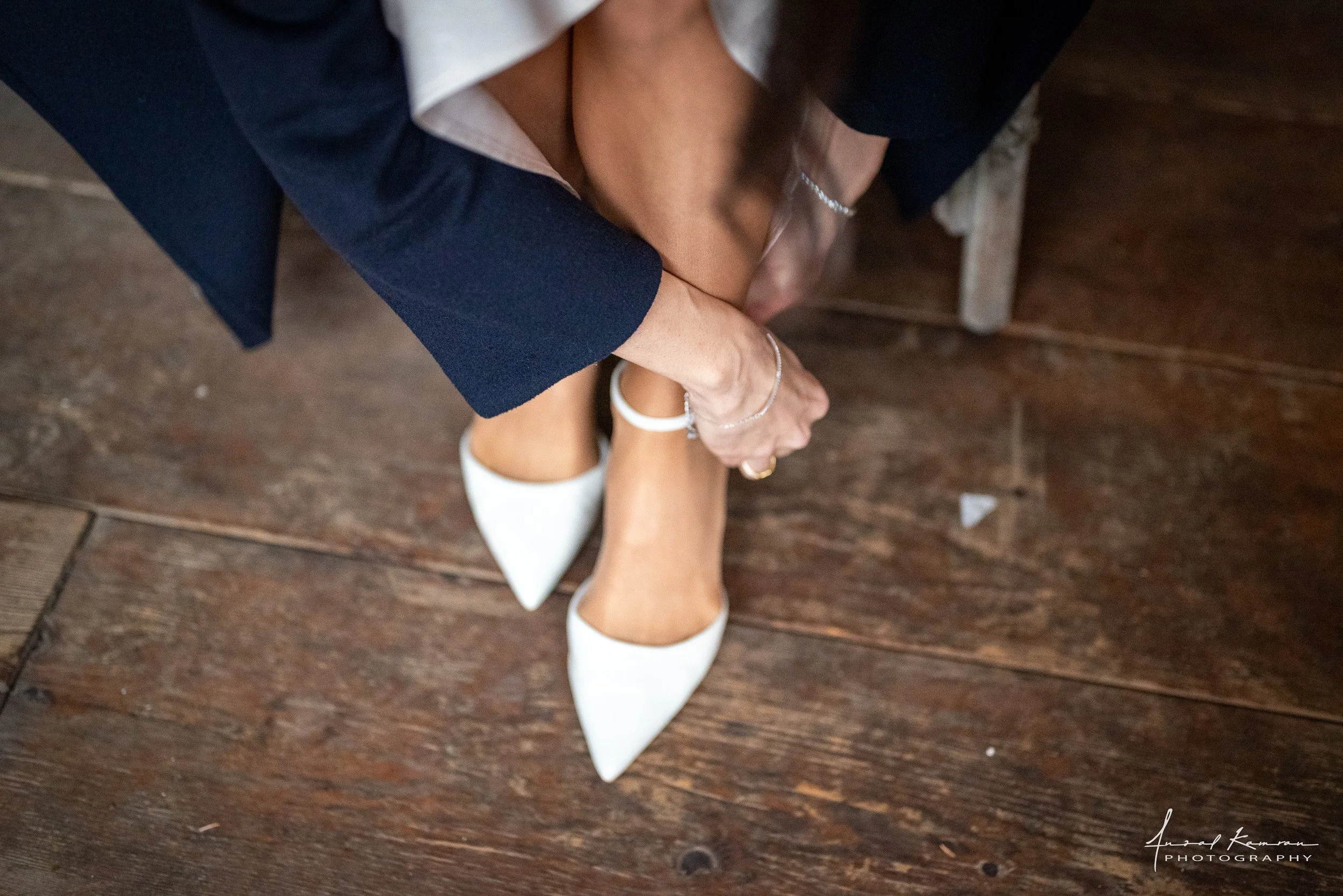A person sitting on a wooden floor, wearing white high heels, a dark blazer, and a white shirt, showing legs and hands with jewelry.