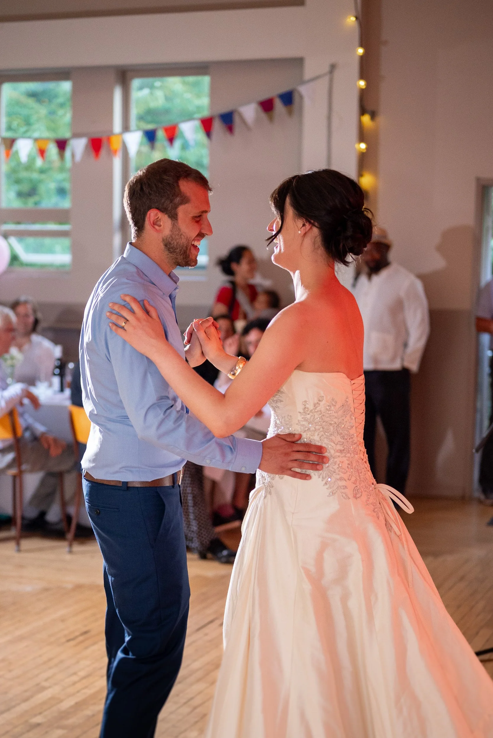 A man in a light blue shirt and dark blue pants dancing with a woman in a strapless wedding dress at a wedding reception. They are smiling and looking at each other, holding hands as they dance. In the background, tables with guests, colorful bunting