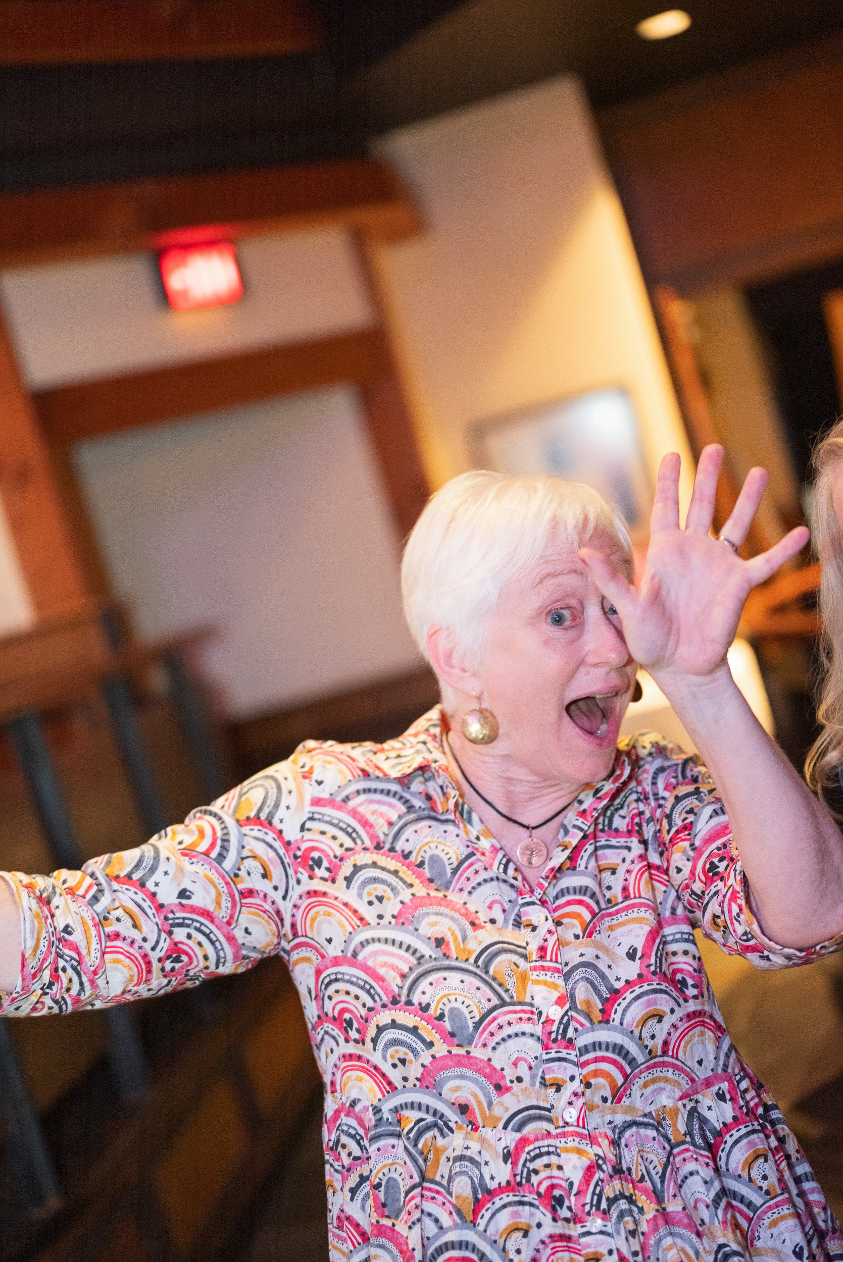 An elderly woman with short white hair and large earrings, wearing a colorful patterned top, is smiling and making a playful gesture with her hand in a lively indoor setting.
