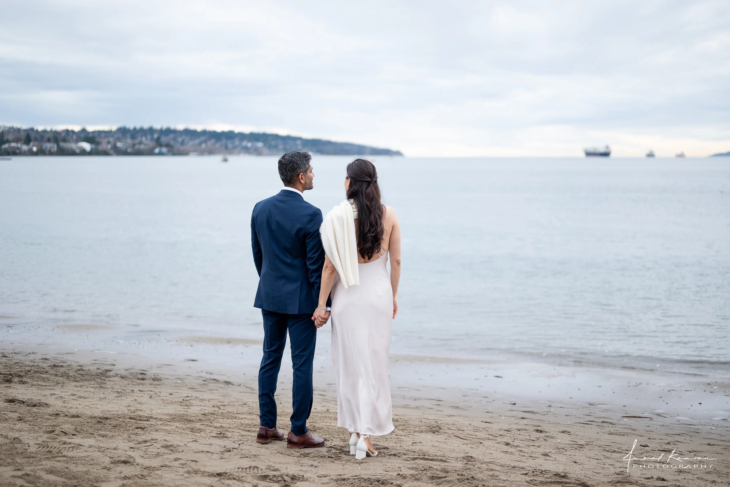A couple dressed in wedding attire holding hands on a beach, facing the water with ships in the background and overcast sky.