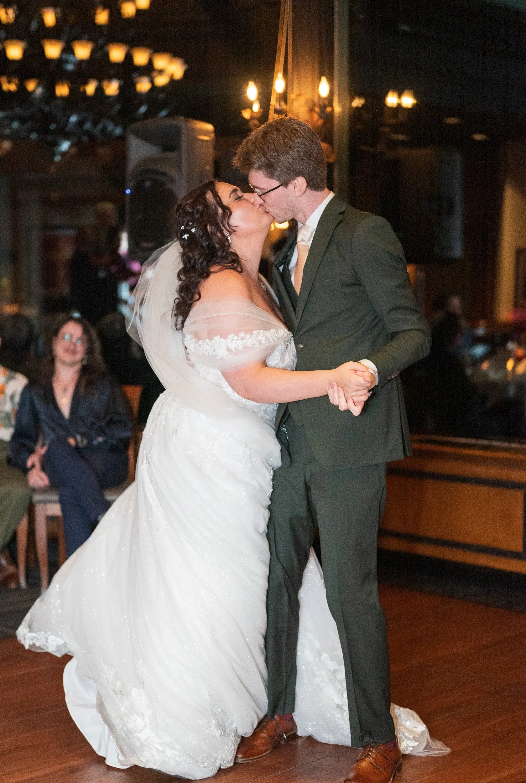 A bride and groom are sharing their first dance at their wedding. The bride is wearing a white gown and the groom is dressed in a dark suit with glasses. They are holding hands and kissing in a warmly lit reception hall.