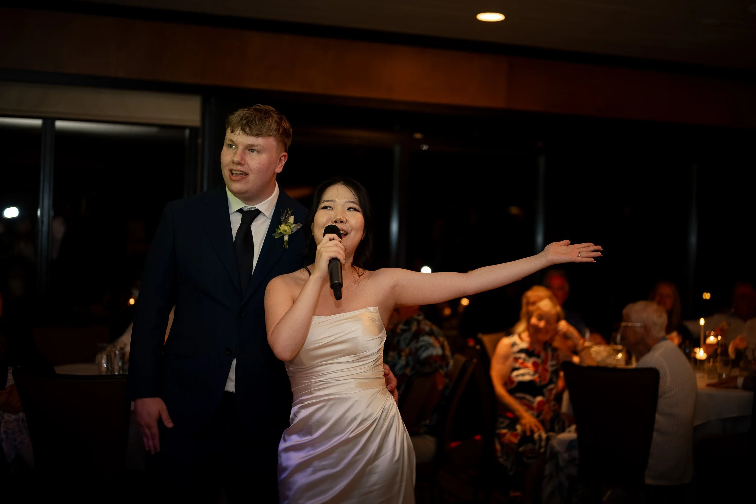 A bride and groom standing together at their wedding reception, with the bride speaking into a microphone and the groom smiling, in a warmly lit indoor venue with seated guests in the background.