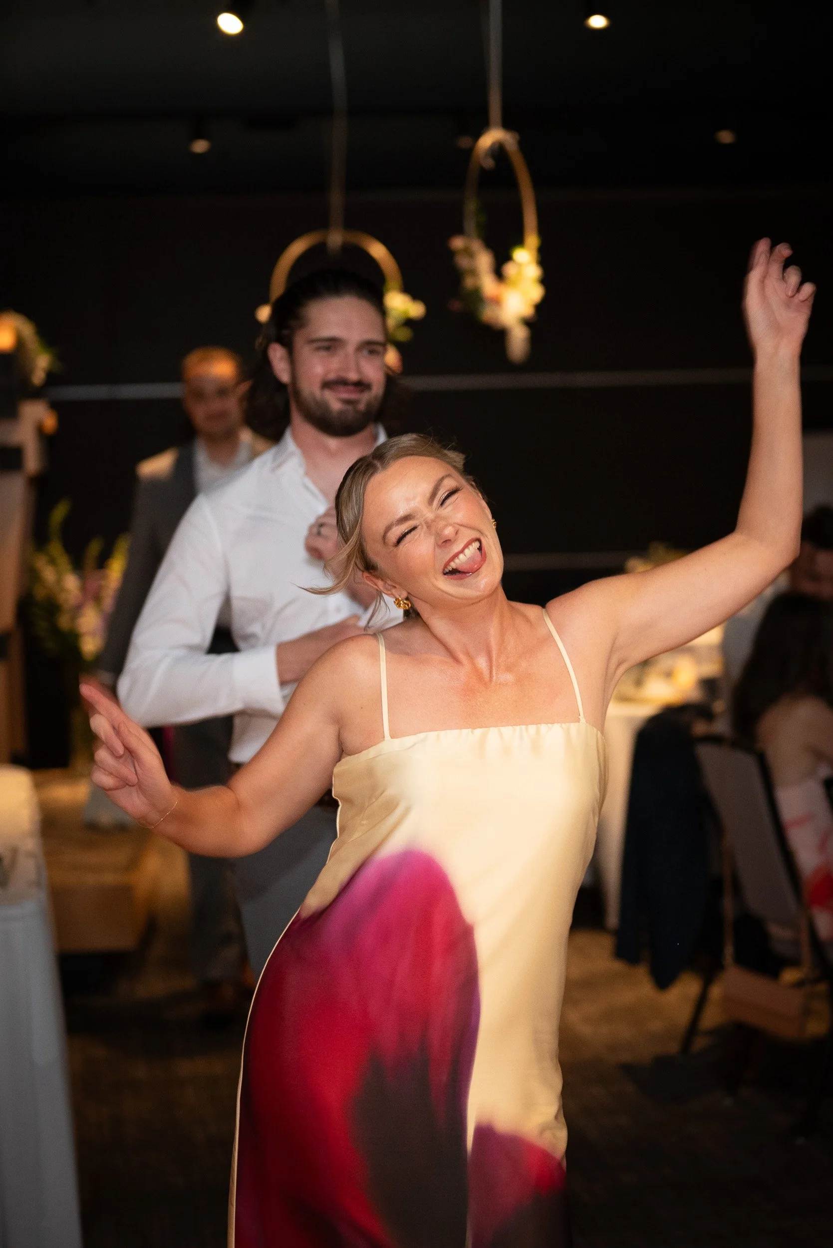 A woman in a yellow dress is dancing joyfully at a celebration, with a big smile and her tongue sticking out, while a man in a white shirt is dancing behind her at an indoor event.