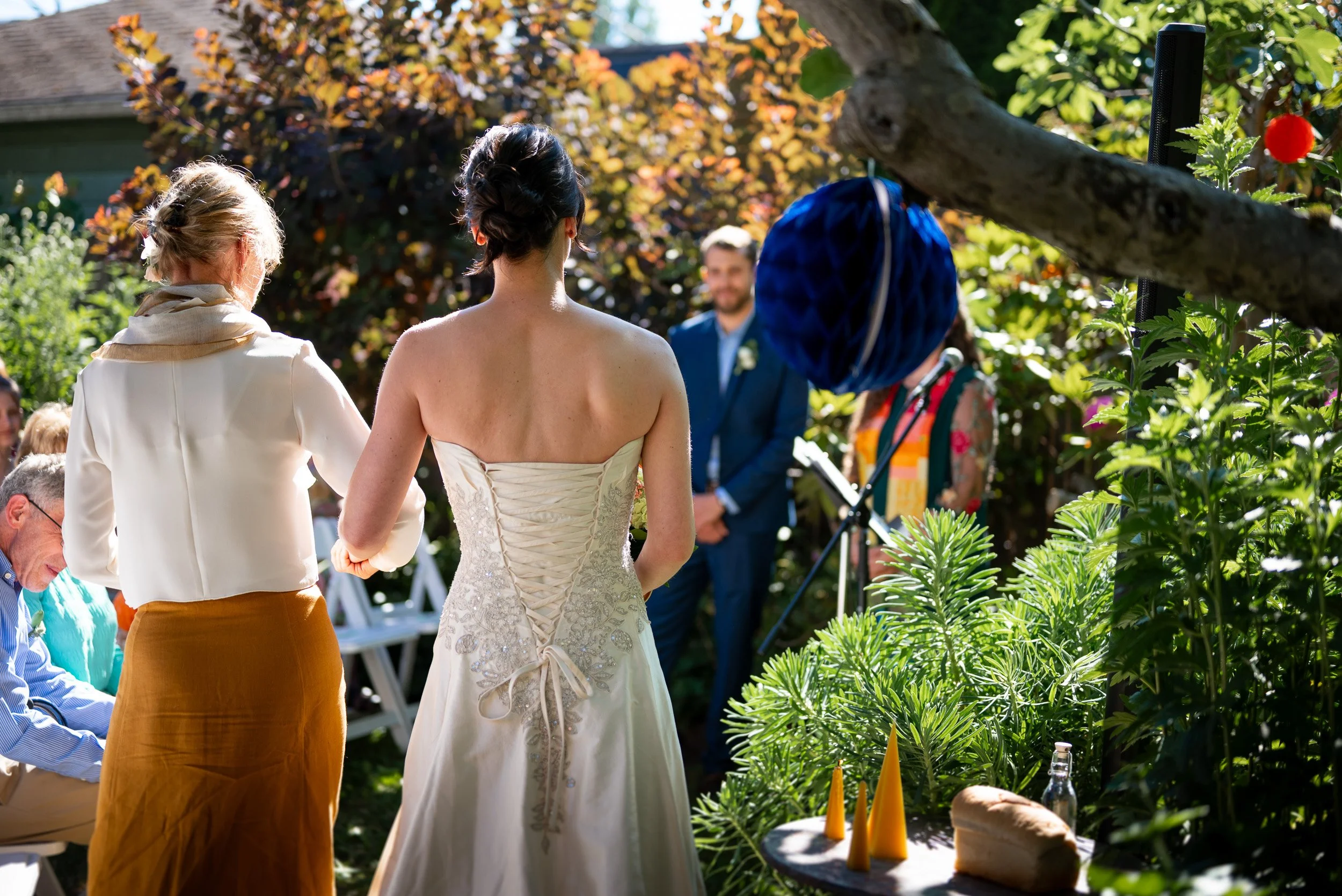 A wedding ceremony outdoors with women in front of the bride, who is wearing a white lace wedding gown, during daytime with sunny weather and lush greenery around.