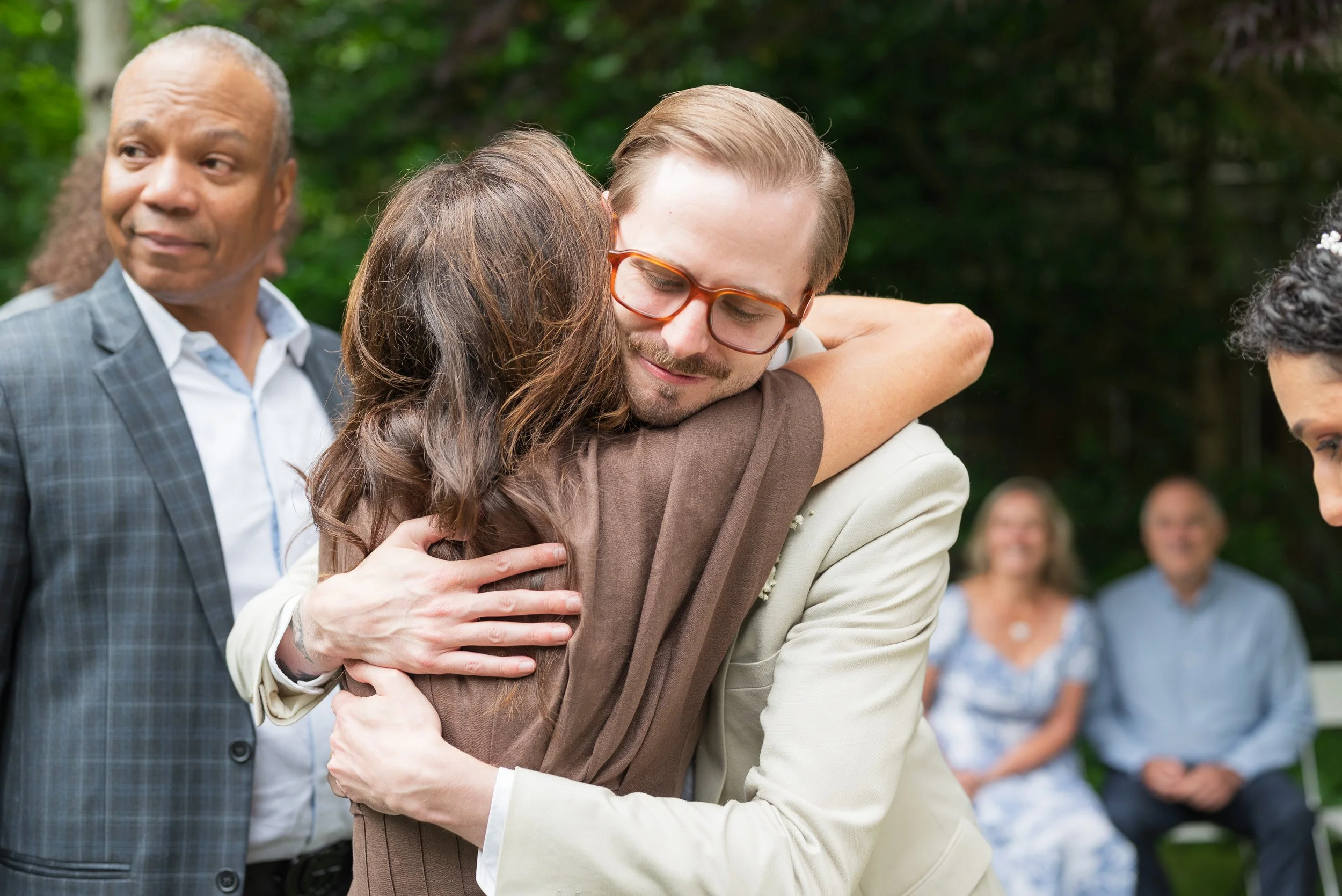 Two people hugging, with one person's face pressed into the other person's shoulder, while others stand in the background outdoors.