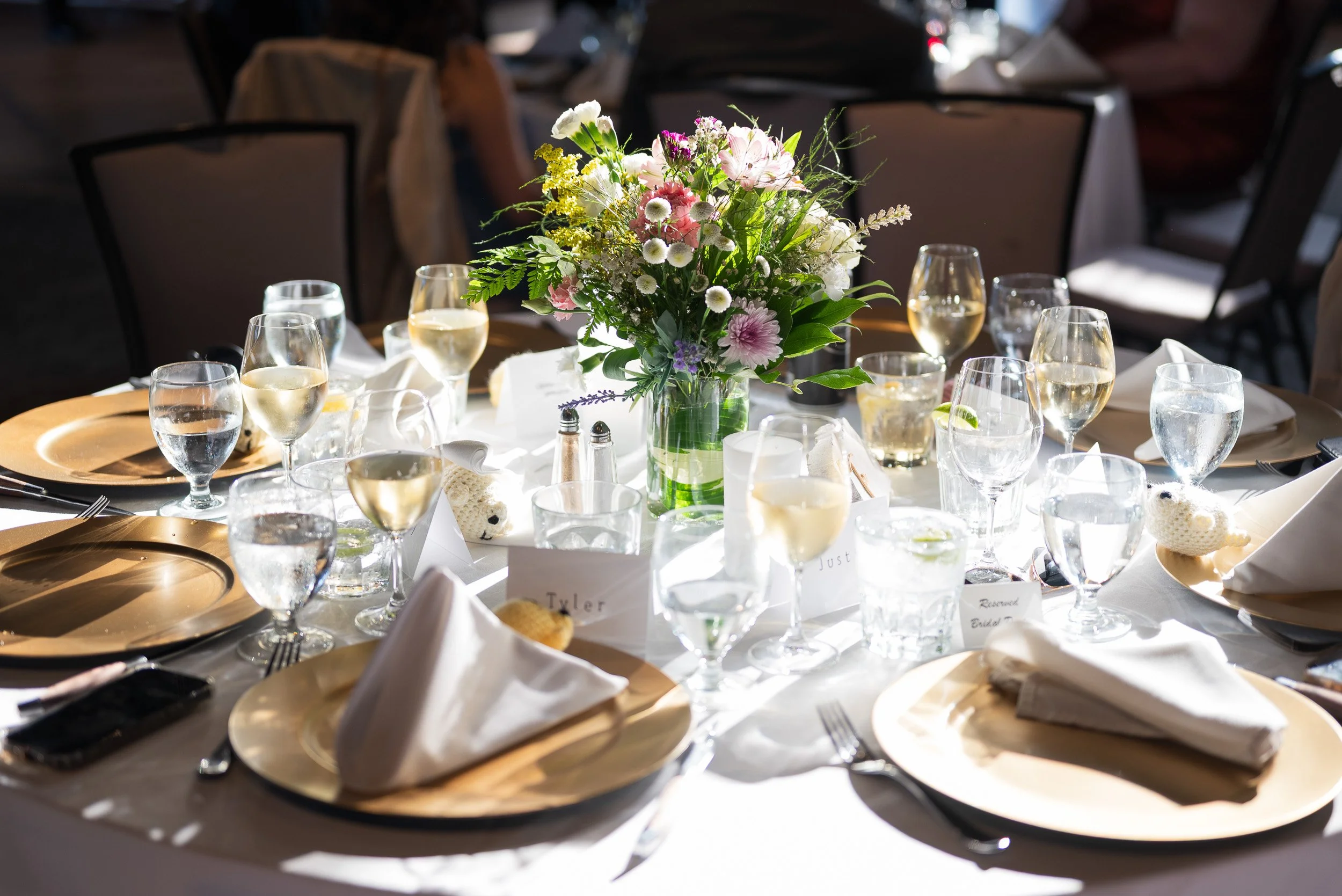A table setup with a floral centerpiece, multiple glasses of water and wine, plates with napkins, and place cards, for a wedding reception at the Seymour Golf and Country Club in North Vancouver.