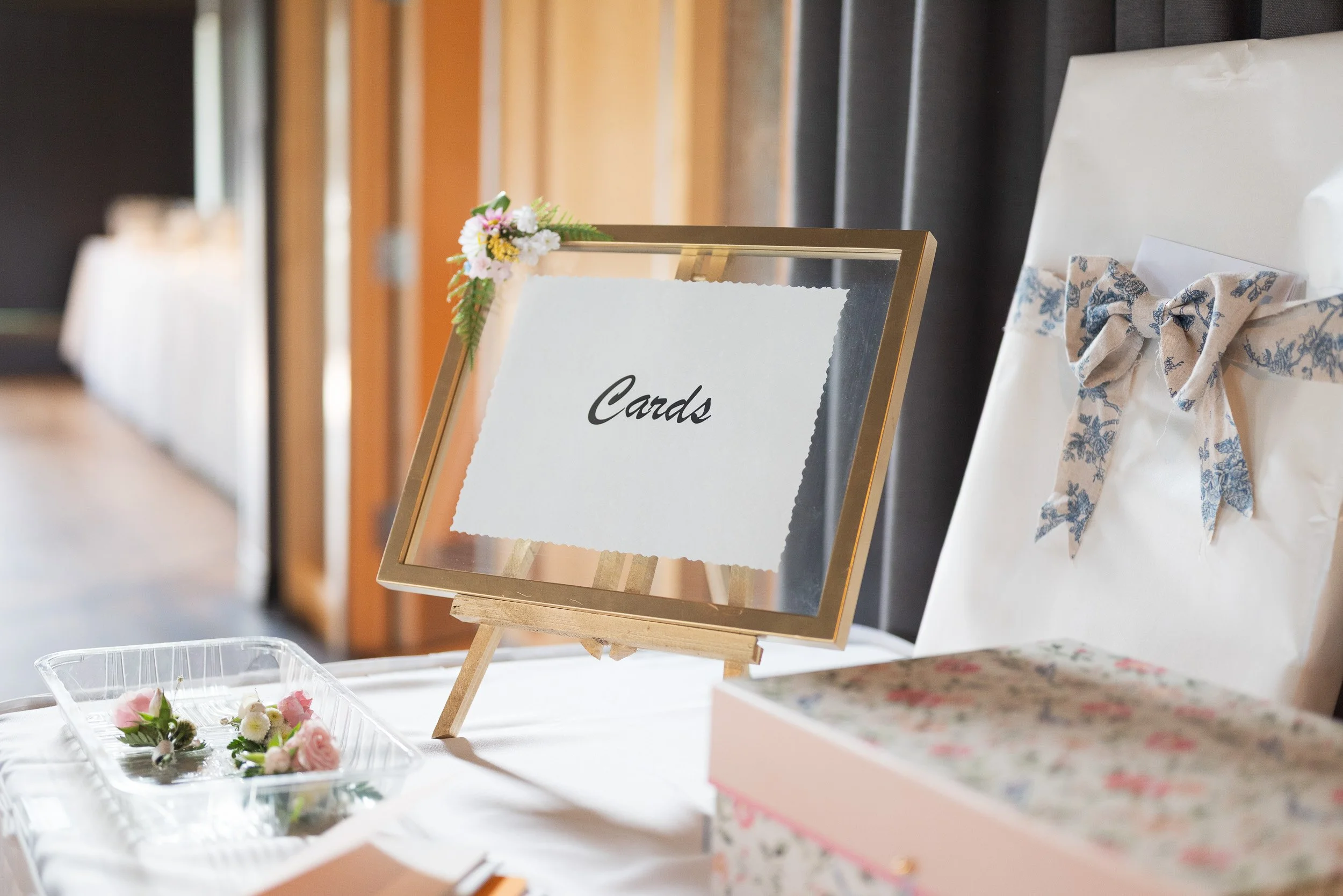 A small wooden easel with a mirror, decorated with pink and white flowers, and a sign that reads 'Cards'. Behind it, there are wrapped gifts with bows, and near the bottom, a plastic container with pink and white flowers.