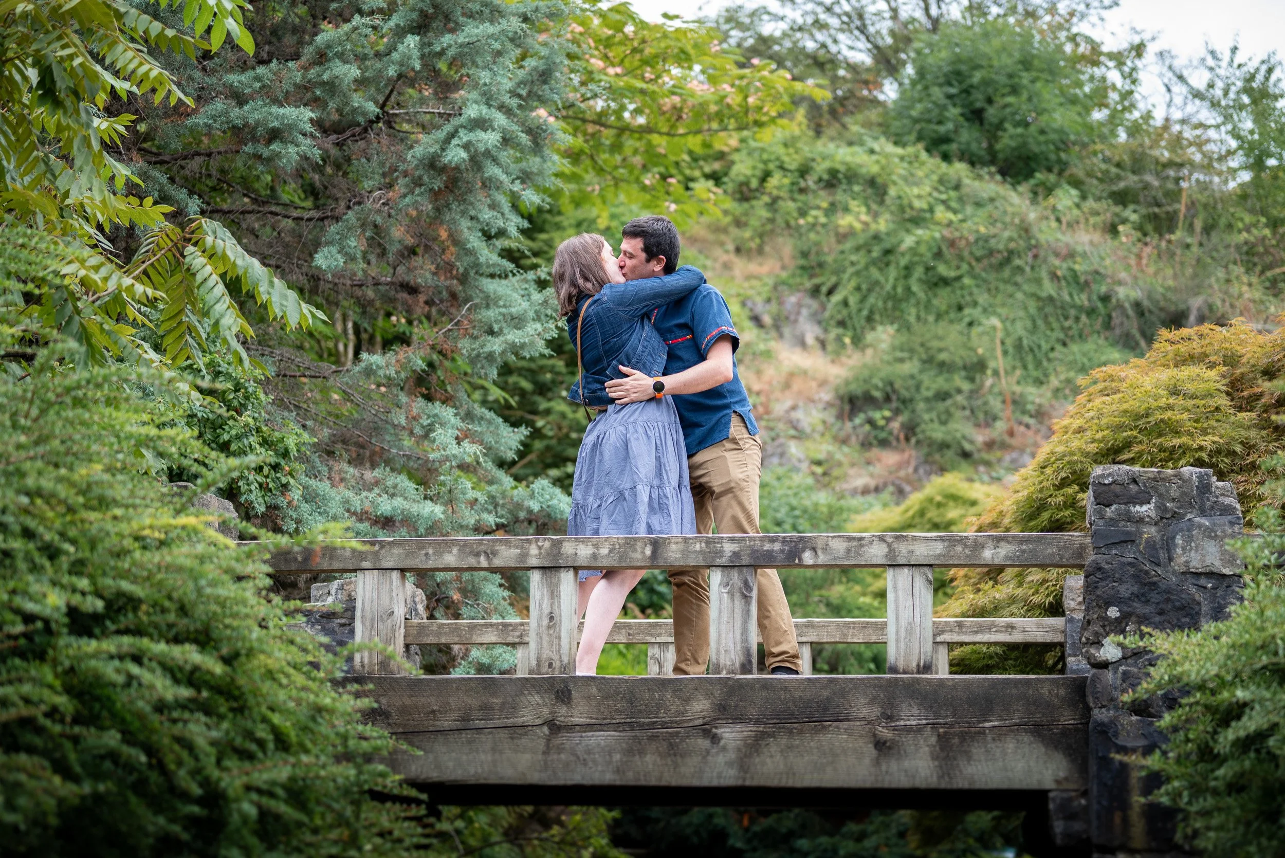 A couple sharing a kiss on a wooden bridge in a lush green park at Queen Elizabeth Park after he proposed.