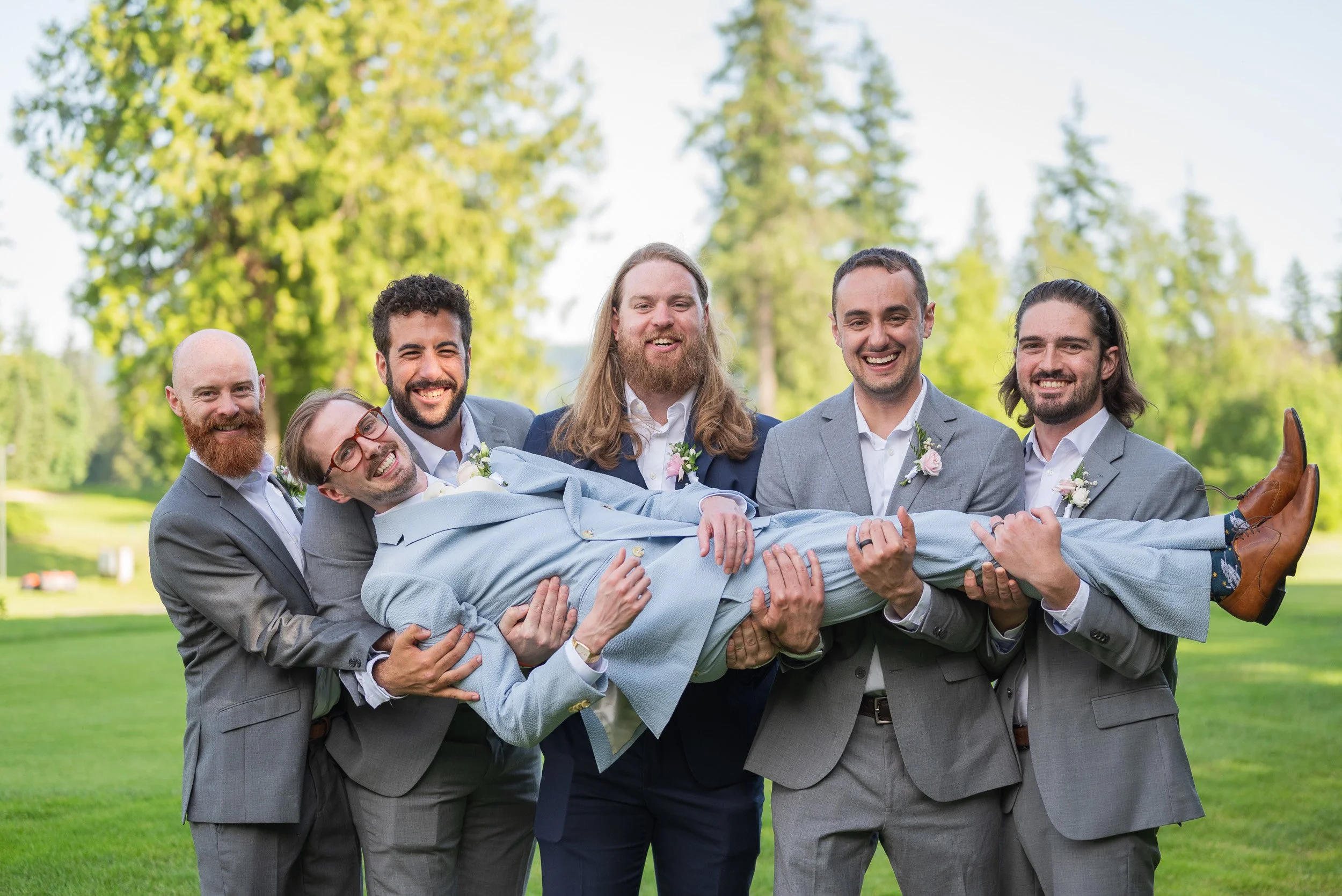 Group of six men in suits holding a man in a blue jacket and colorful socks in a park with green trees in the background.
