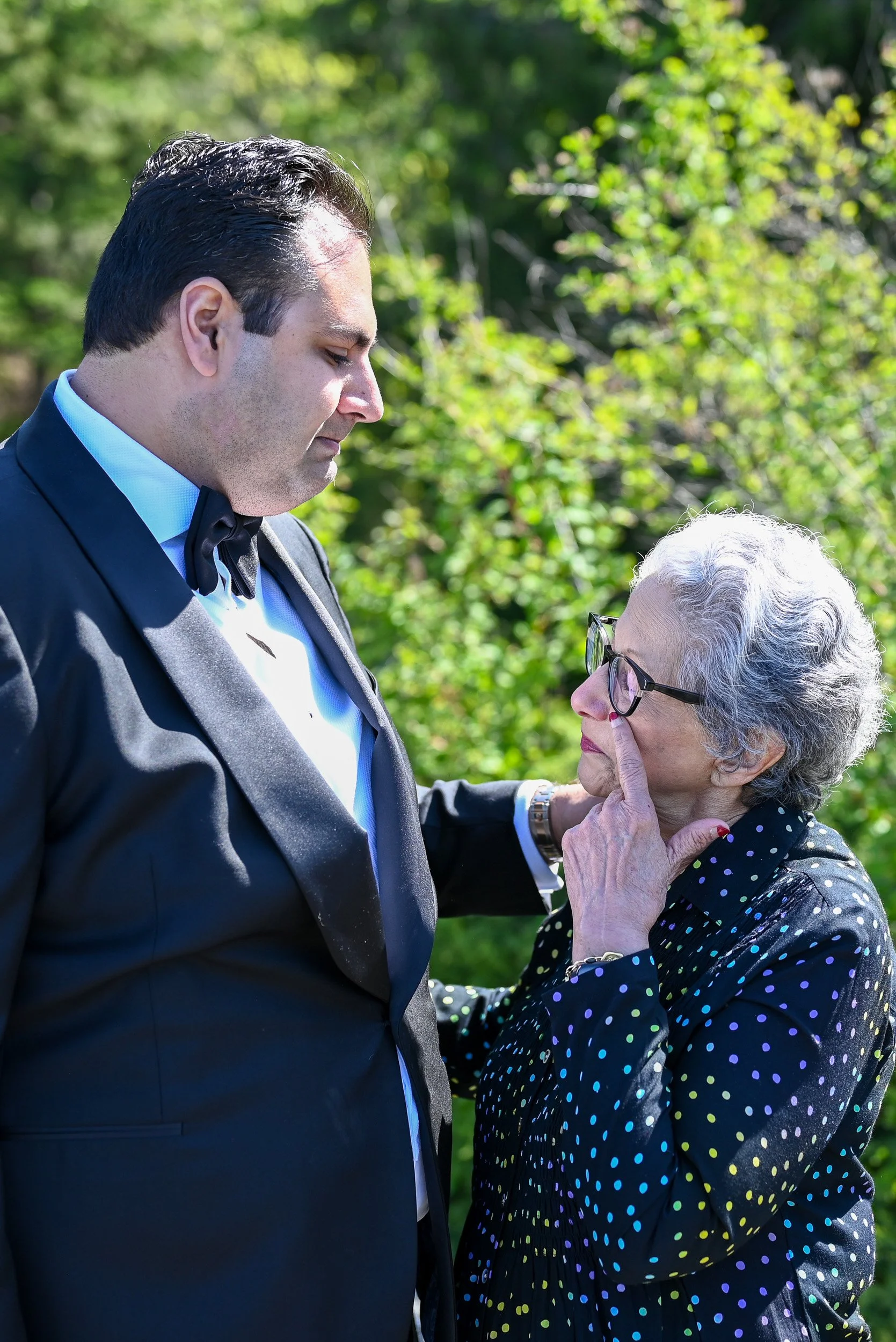 A man in a formal tuxedo with a bow tie standing outdoors with an elderly woman who is touching his face gently. The woman has gray hair, glasses, and is wearing a polka dot blouse. Green foliage is in the background.