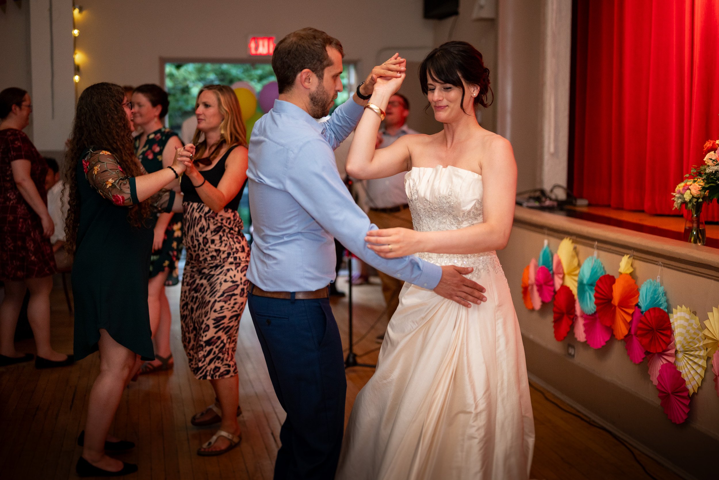 A couple dancing at a wedding reception, surrounded by friends, with colorful paper decorations on the wall and balloons in the background.