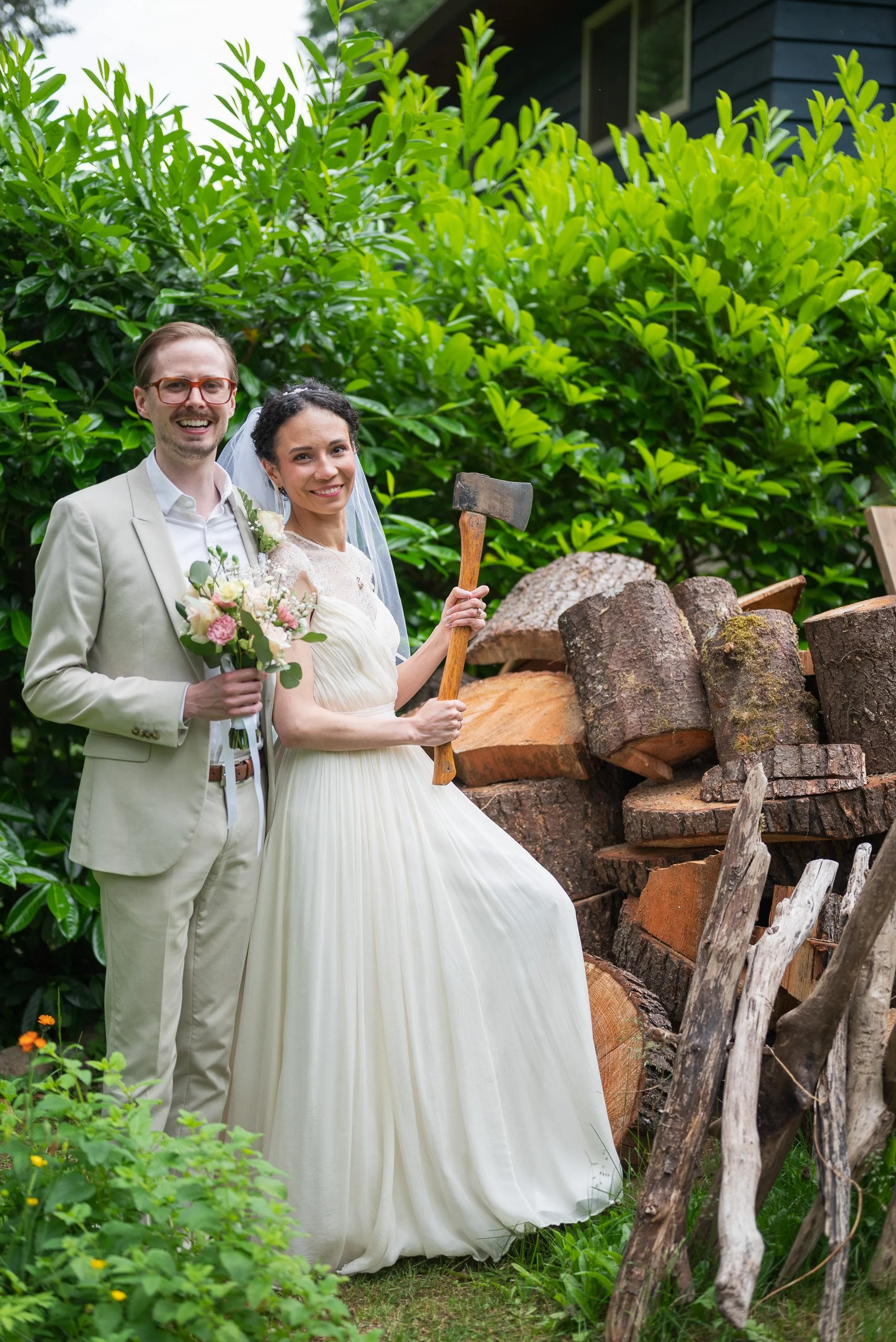 A smiling bride and groom in wedding attire standing outdoors among green bushes and logs, with the bride holding a small bouquet and an axe.