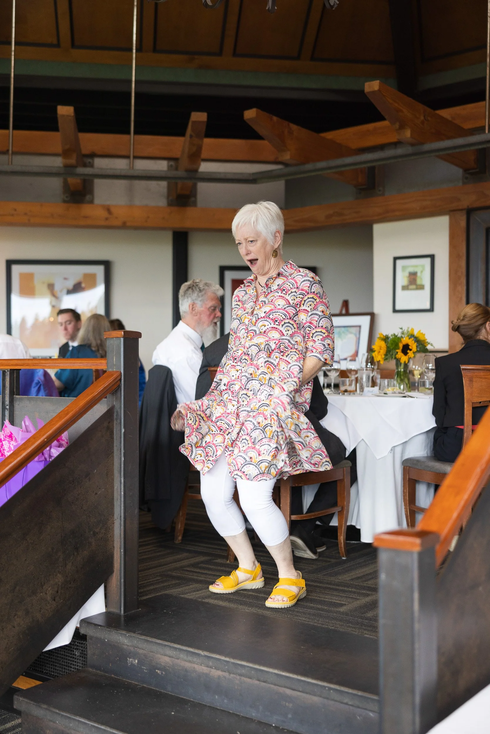 A woman with short white hair wearing a colorful patterned dress with puffed sleeves, white leggings, and yellow sandals standing on a black staircase inside a restaurant or event hall. She appears to be surprised or reacting to something. There are 