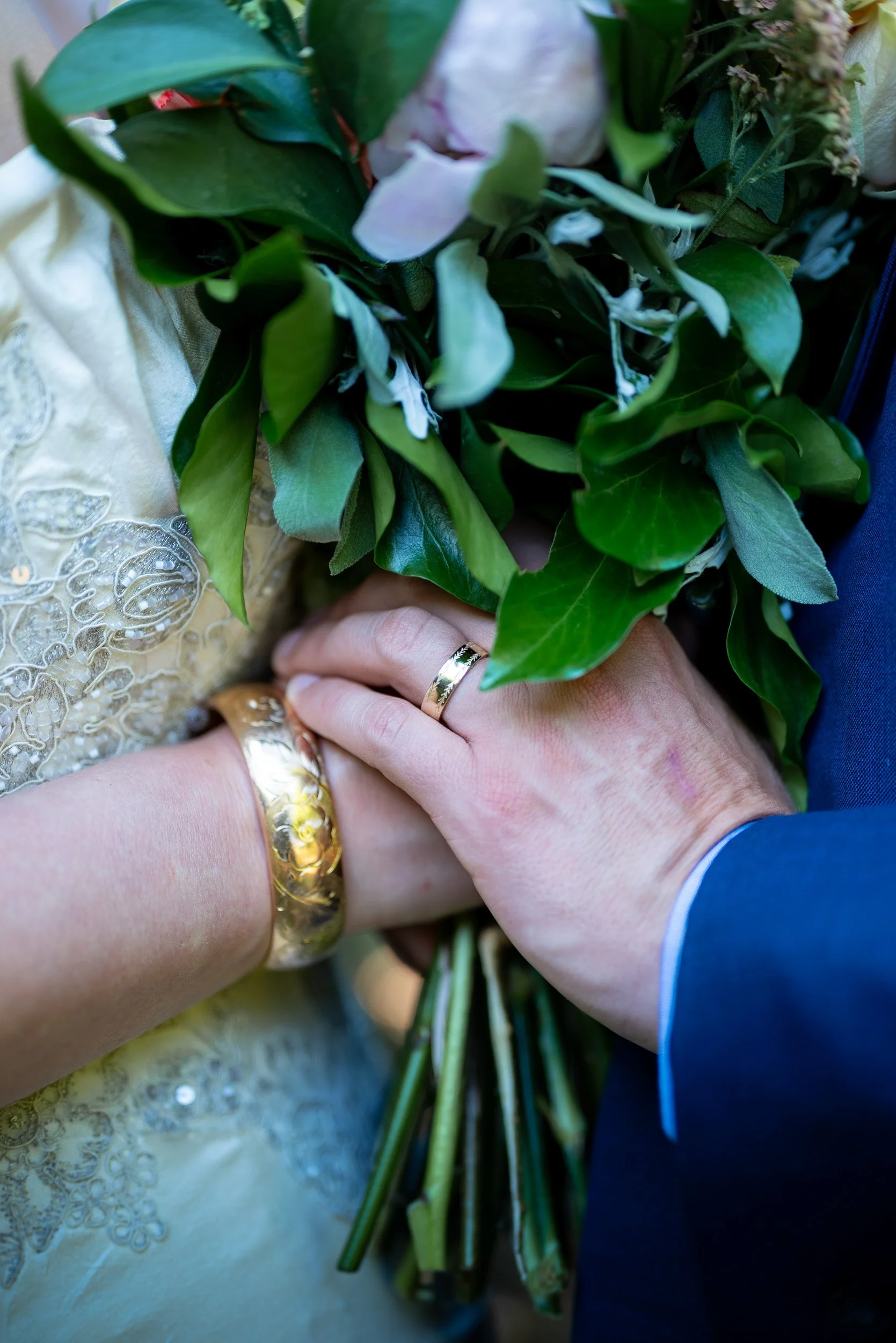 Close-up of hands with wedding rings holding a bouquet of green leaves and flowers, person wearing an embroidered gold dress and a blue suit.