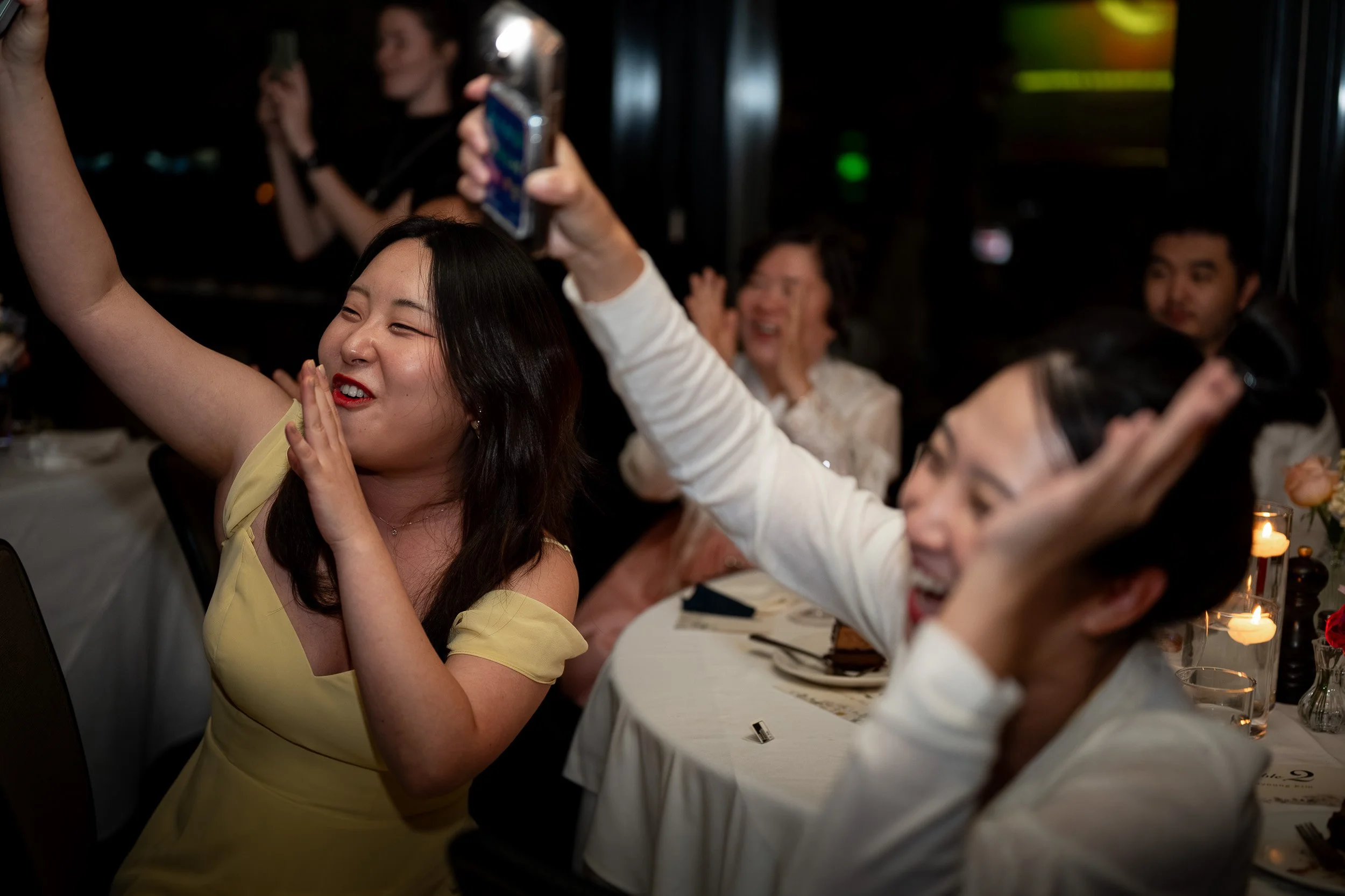 A group of people at a table, celebrating and taking selfies at a party or dinner event in a dimly lit setting.