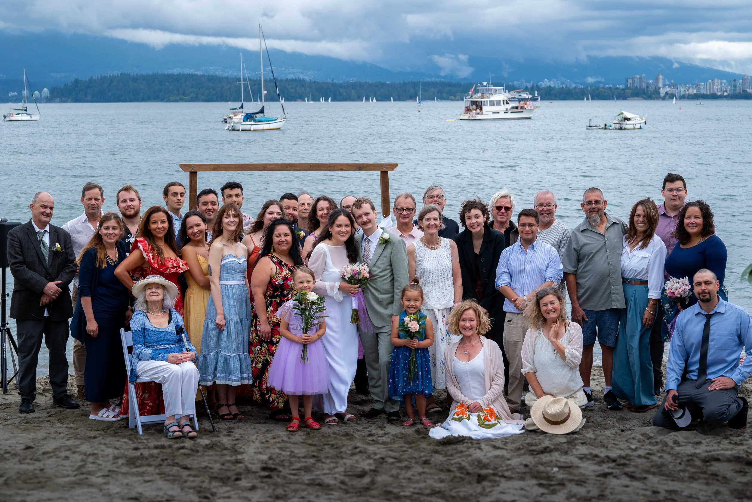 A large group of people gathered on a beach for a wedding at Jericho beach Vancouver, with sailboats on the water and a city skyline with mountains in the background.