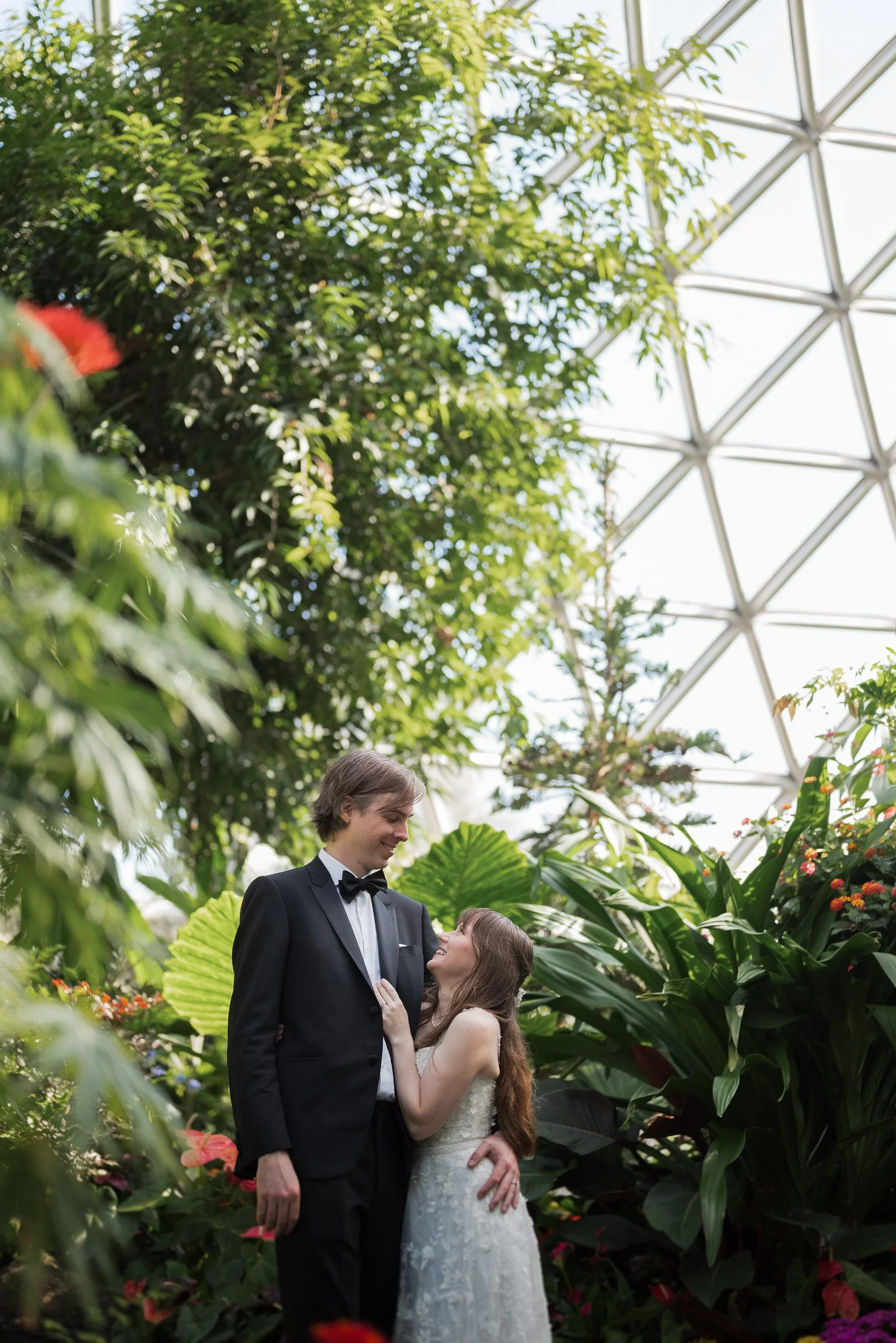 A bride and groom sharing a moment in a lush botanical setting at Bloedel Conservatory, with the groom looking at the bride and smiling, surrounded by green plants and flowers, under a glass domed ceiling.
