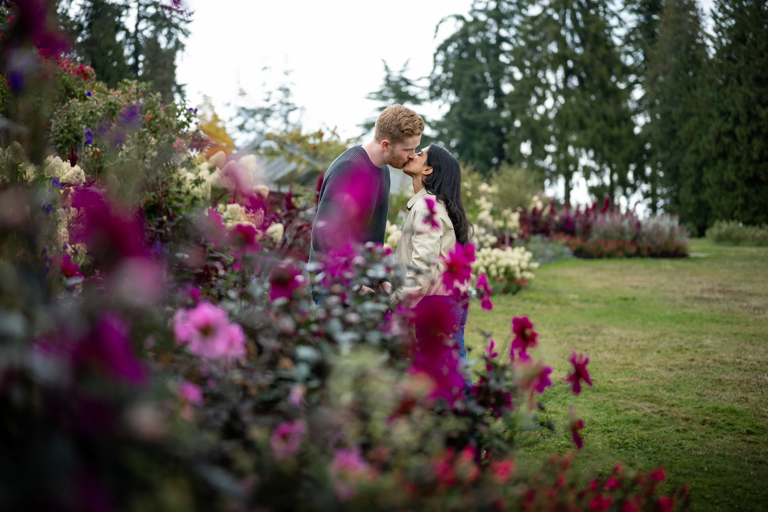 A couple sharing a kiss amidst a garden of colorful flowers and trees in the background at Stanley park rose garden engagement.