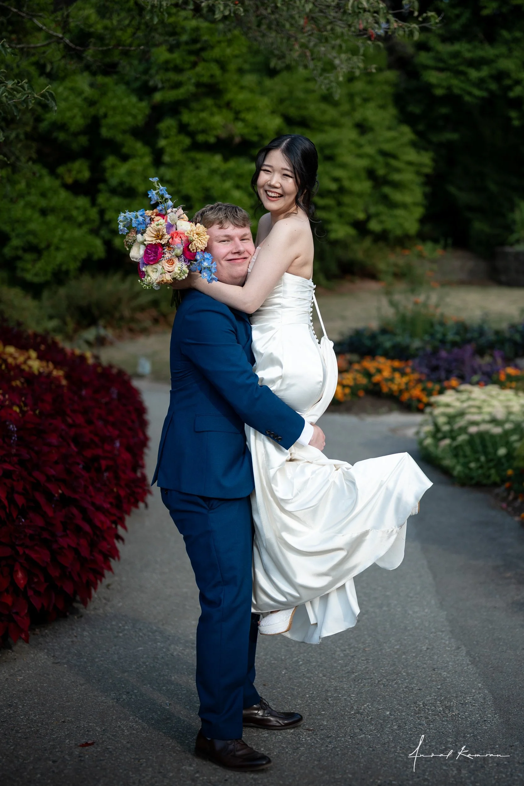 A man in a blue suit lifting a woman in a white wedding dress, who is holding a colorful bouquet, outdoors with green trees and flowers at Queen Elizabeth Park.